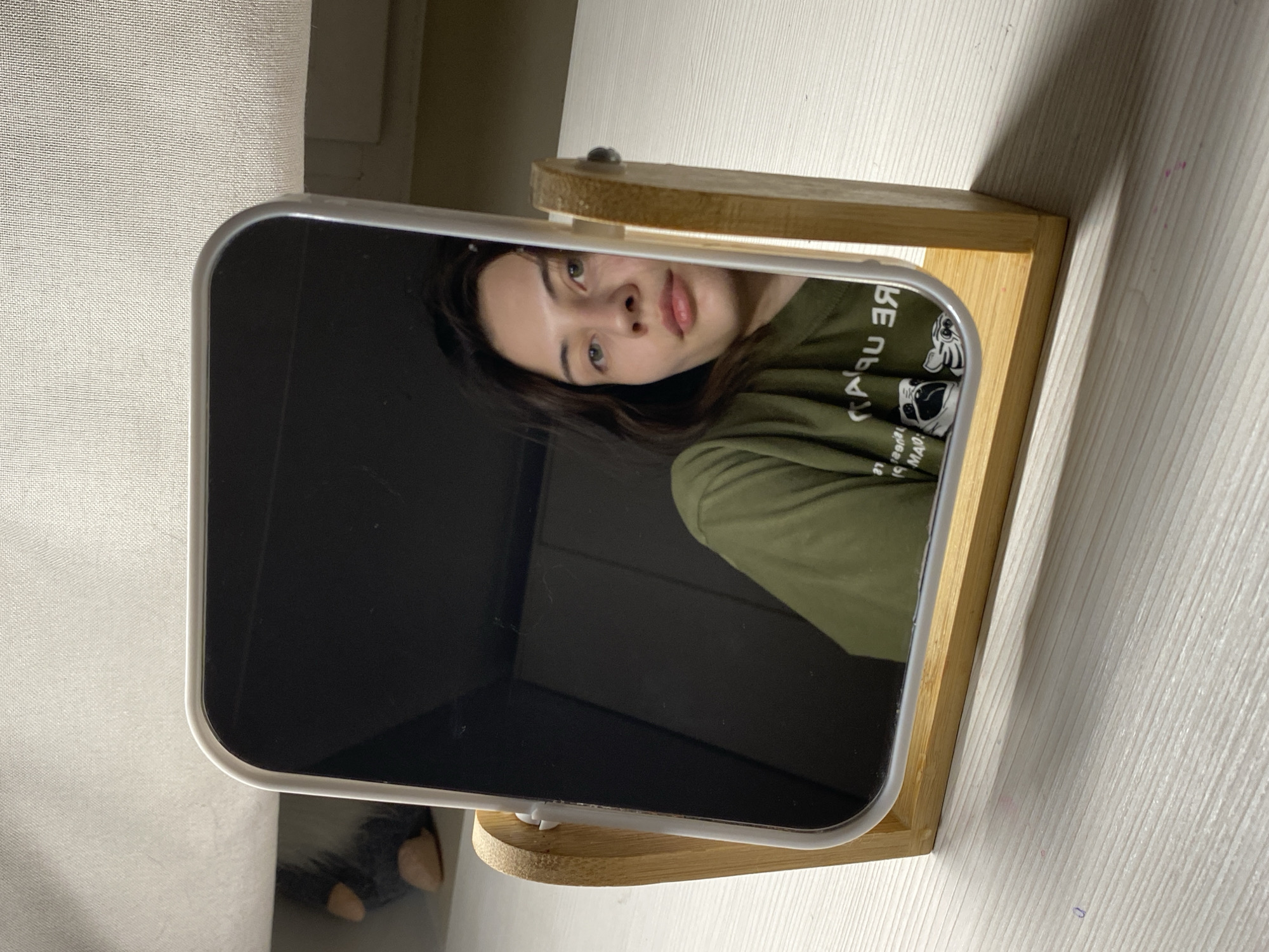 A young woman with dark hair and light-colored eyes (greenish-hazel) is visible in the reflection of a rectangular mirror. She has a neutral expression and appears to be wearing an olive green long-sleeved shirt. On her shirt, visible as mirrored text, are the words "BE ПРИ" and beneath that, "МОЙ МАД," surrounding a graphic of a pug dog wearing glasses.
The mirror has rounded corners and a sleek, light-colored frame, possibly made of metal or plastic. It stands upright on a light-colored wooden base or stand, which has a natural grain. To the left of the mirror, a light-colored, textured surface resembling a curtain or fabric wall covering is visible. To the right, the mirror and its stand rest on a light-colored wooden surface, likely a desk or table, which also shows a subtle wood grain. Shadows are cast on this surface, suggesting indoor lighting, possibly from a window to the left. A small, dark object, possibly a plush toy or part of a shoe, is partially visible at the bottom left, outside the mirror's reflection. The scene depicts an indoor setting, likely in Votkinsk, Russia.