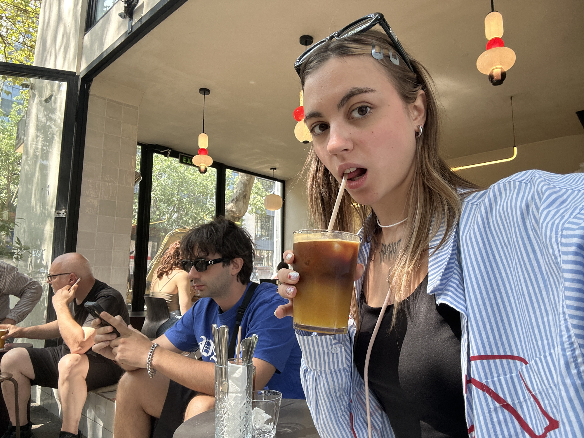 The image depicts a group of people at an outdoor cafe in Tbilisi, Georgia. In the foreground, a young woman with blonde hair and glasses is holding a glass of iced coffee and looking towards the camera. She is wearing a blue and white striped shirt over a black tank top. To her left, a man with dark hair and sunglasses is seated, holding a smartphone. Further to the left, an older man with glasses and a shaved head is also seated, holding a cigarette. The cafe appears to be situated near trees and buildings, suggesting an urban setting. The overall ambiance suggests a casual gathering on a sunny day.