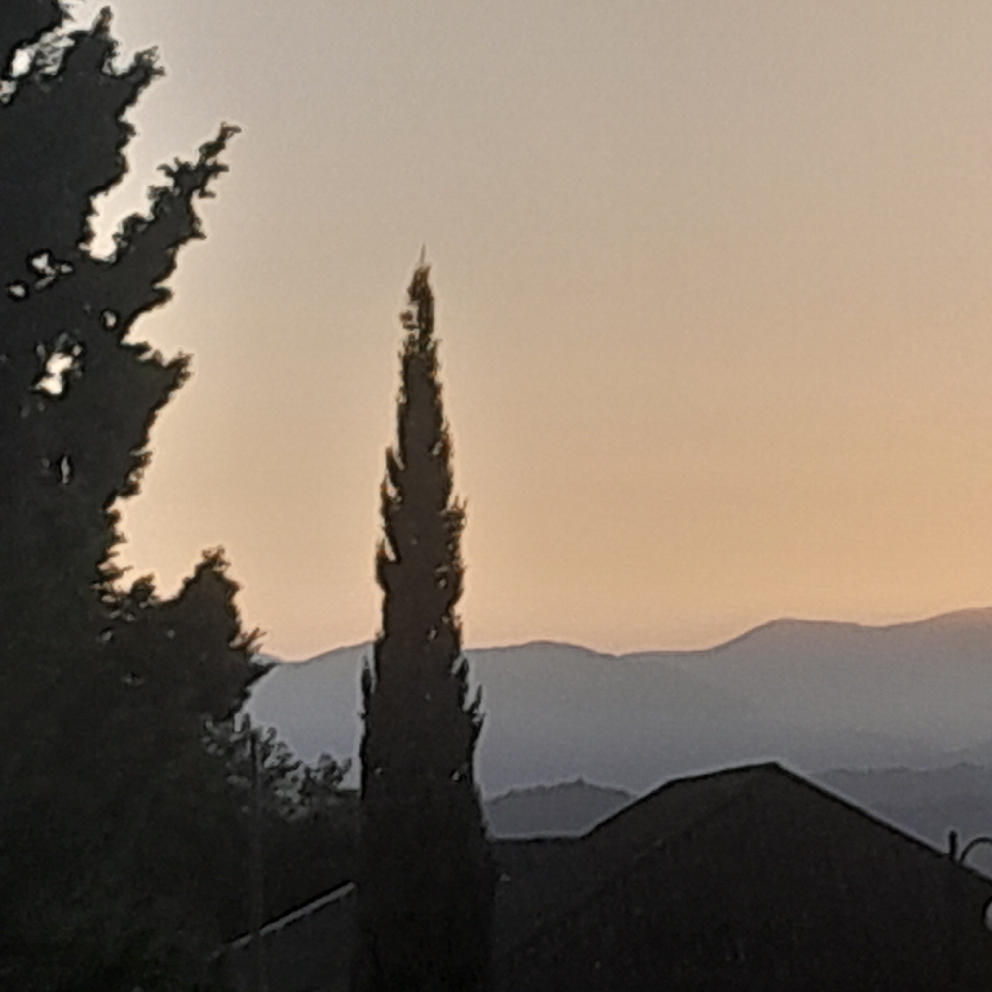 The image depicts a silhouette of a tall, slender tree in the foreground, possibly a cypress, against a soft, gradient sky of pale orange and beige, indicative of either dawn or dusk. In the mid-ground, a range of hazy blue mountains forms a layered backdrop, suggesting a landscape view. At the very bottom of the frame, the dark silhouette of a building's roof is visible, with a curved element at its right edge that could be part of an antenna or architectural feature. The overall scene is peaceful and serene, with no people or animals visible. The setting appears to be a natural landscape near a populated area, possibly in a city like Astara, Azerbaijan, known for its proximity to both the Caspian Sea and mountainous terrain.