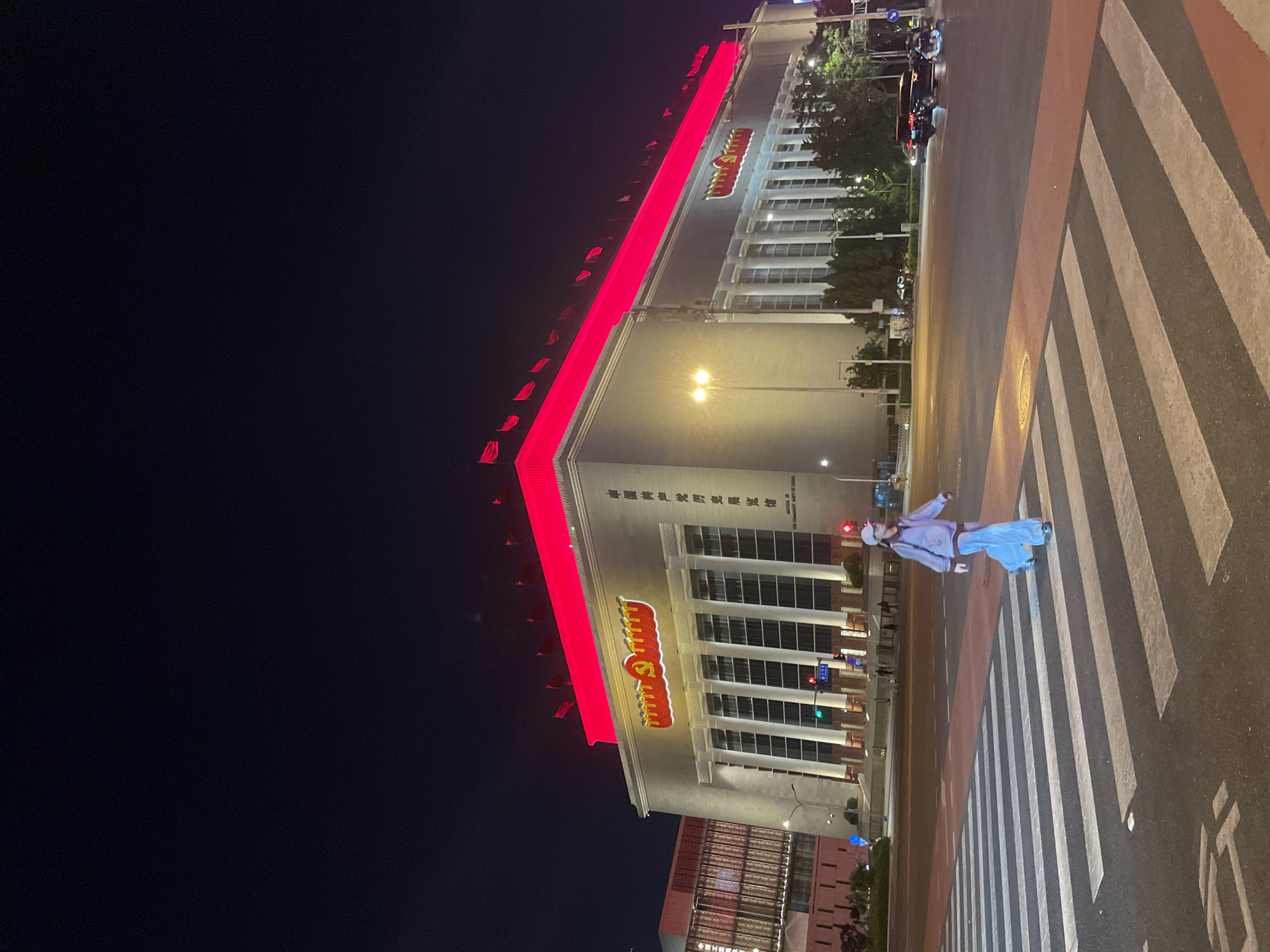 The image depicts a nighttime scene in Chaoyang District, China. A pedestrian is crossing a zebra stripe crosswalk. Behind the person is a large building illuminated with red lights, and the Chinese Communist Party symbol is visible. The sky is dark.
