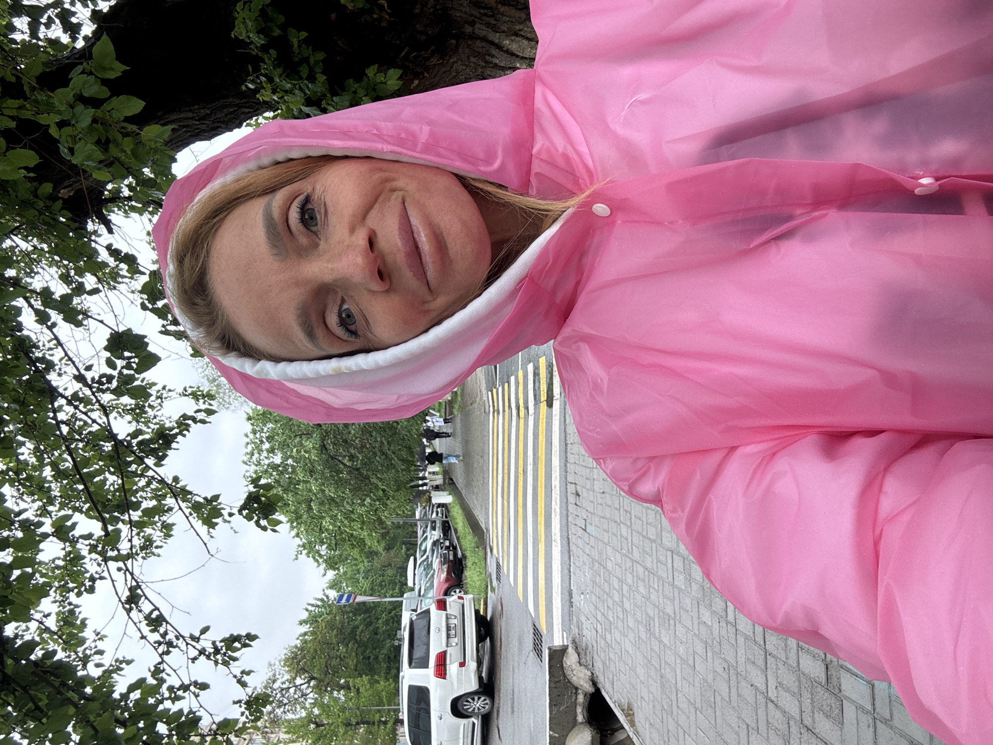 A woman wearing a vibrant, somewhat translucent pink rain poncho with a white-lined hood is featured in a close-up, portrait-style shot. She has blonde hair partially visible under the hood and light-colored eyes, looking towards the camera with a calm, subtle smile.
The background reveals an urban street scene under an overcast daytime sky. On the left, lush green tree branches and a dark tree trunk frame the upper portion of the image. On the right, a paved sidewalk and street are visible, featuring a crosswalk with distinct yellow and white stripes. A line of parked cars extends along the street, with a white SUV prominently in view. The SUV's rear license plate is partially readable, showing "KG 01 B," which corresponds to Bishkek, Kyrgyzstan, as per the location context. A street sign can be seen further down the road.
The woman's rain gear suggests either recent or impending wet weather. The scene captures a casual moment on an ordinary city street.