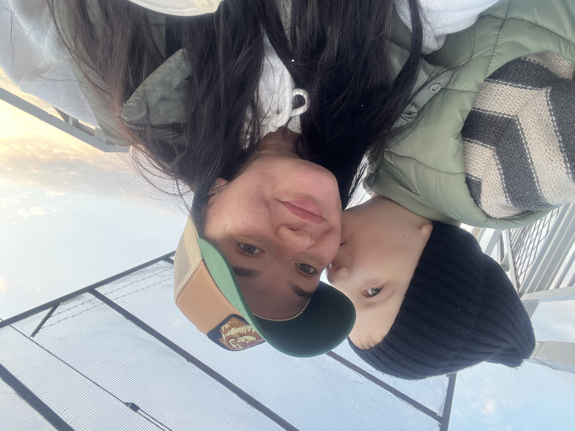 A selfie-style, slightly upside-down photo captures a woman and child. The woman, wearing a green and tan baseball cap with a patch on the front, looks directly at the camera. A child in a black knit cap is close to the woman's face, appearing to be kissing her cheek. The woman is wearing a white hoodie and a light green puffer jacket, with a patterned scarf around her neck. Behind them is a wire mesh fence and a sky with pale clouds. The location might be an elevated platform or rooftop in Kazan, Russia, though the specific details needed to confirm this are not visually evident in the image.