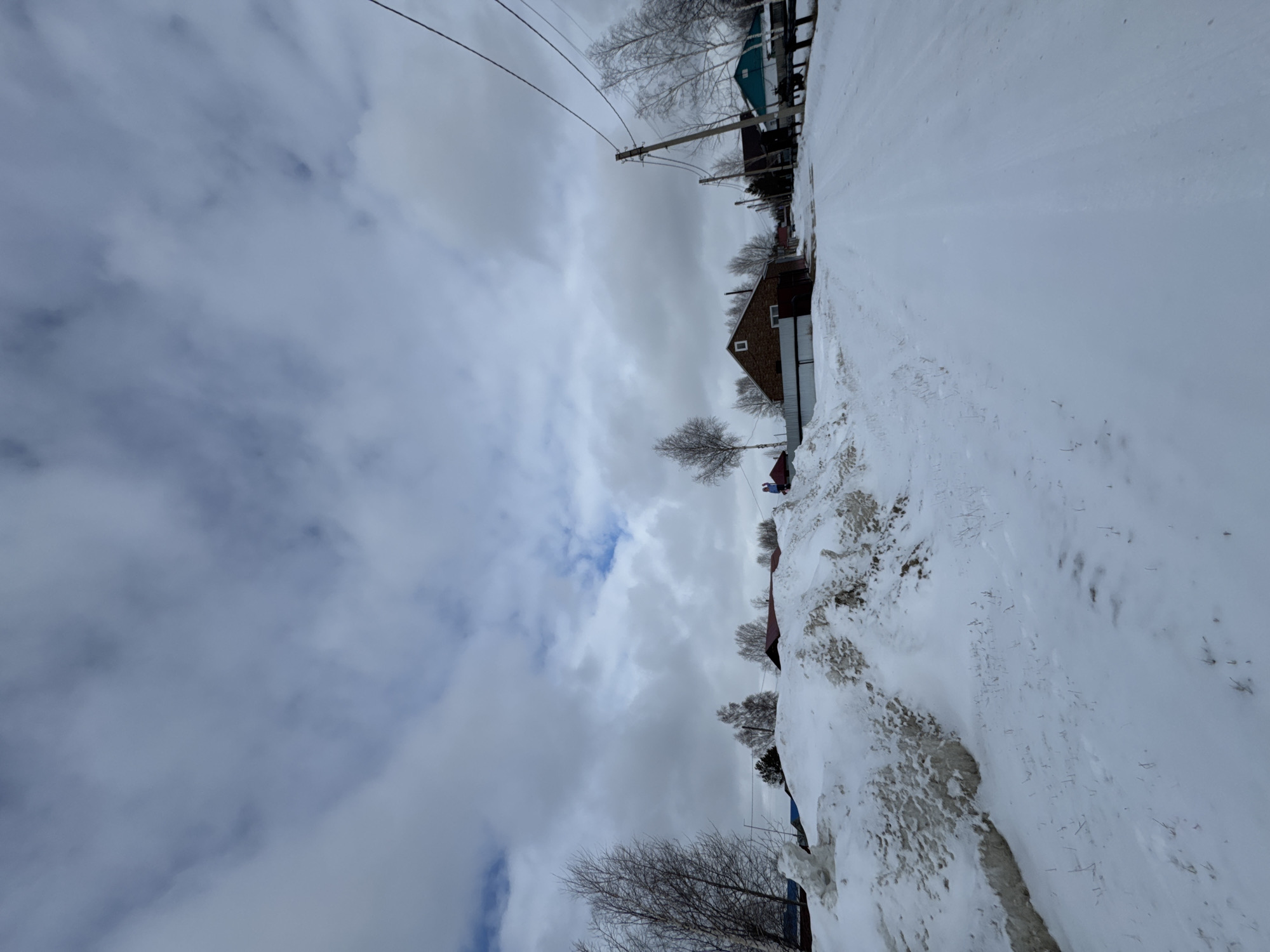 The image shows a winter scene in Высокий, Russia. Snow covers the ground, obscuring the edges of a road. Several small buildings are present on the right, including at least one house constructed of red brick. Overhead powerlines run in the distance, along with thin, leafless trees. The sky is primarily covered with grey clouds. A figure wearing a red hat is present between buildings.