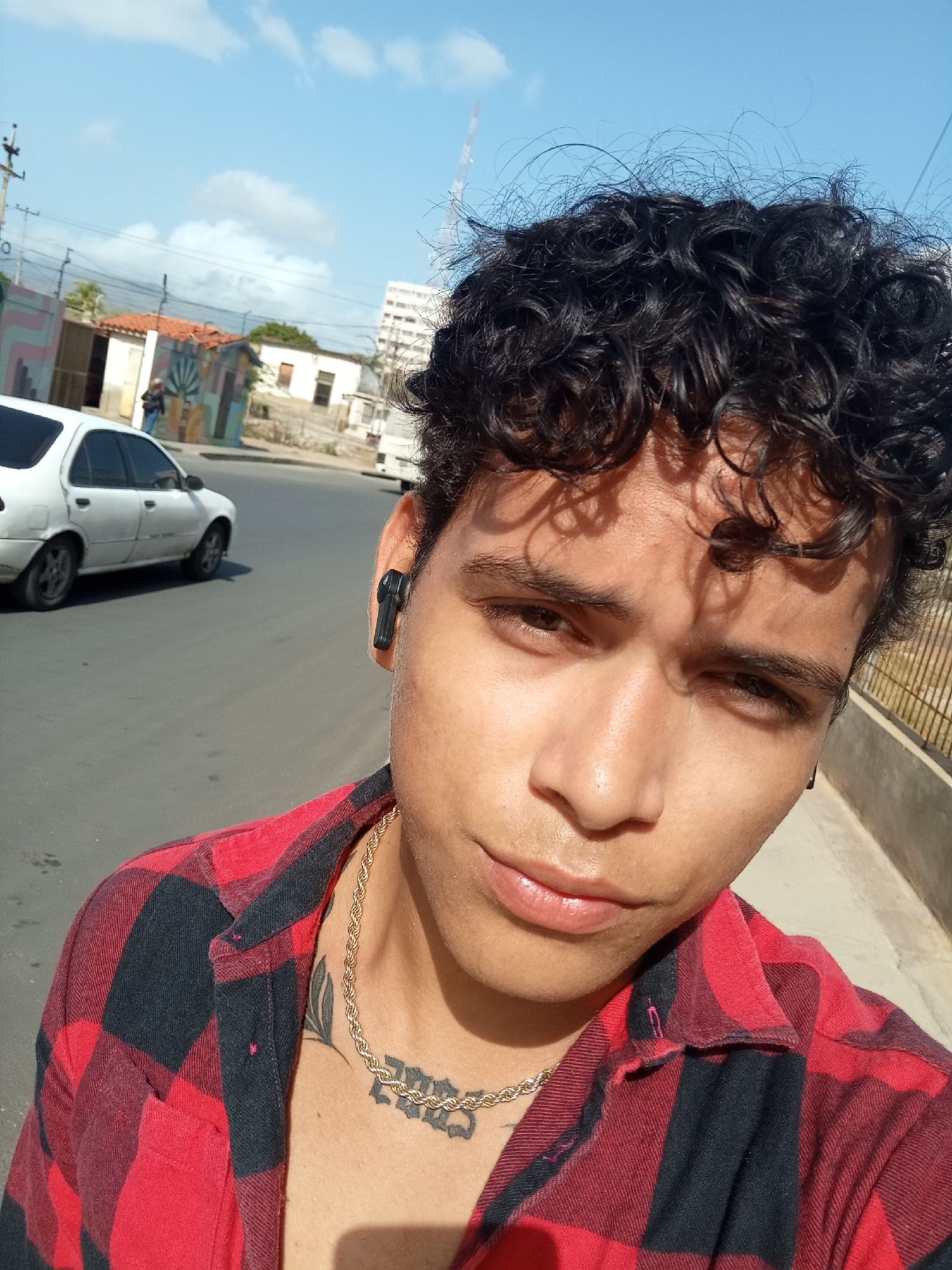 A young man is captured in a close-up selfie taken outdoors on a bright, sunny day, likely in Juan Griego, Venezuela. He appears to be in his late teens or early twenties, with dark, curly hair and brown eyes. He wears a red and black plaid shirt, unbuttoned to reveal a gold chain necklace and two tattoos on his chest: one that appears to be stylized text in a script or gothic font, and another resembling a leaf or abstract design. A black wireless earbud is visible in his right ear. His expression is neutral as he looks directly into the camera.
The background shows an urban or semi-urban street scene. To the left, a white four-door sedan is visible on the road, with a utility pole and power lines above it. Further back, several residential buildings of varying styles and colors can be seen, some featuring murals. One building has a distinct red-tiled roof. A partial view of a white truck and a metal fence is also visible on the right side of the image. The sky is clear blue with sparse white clouds, and the strong sunlight casts shadows on the man's face, particularly from his hair. No legible text is visible.