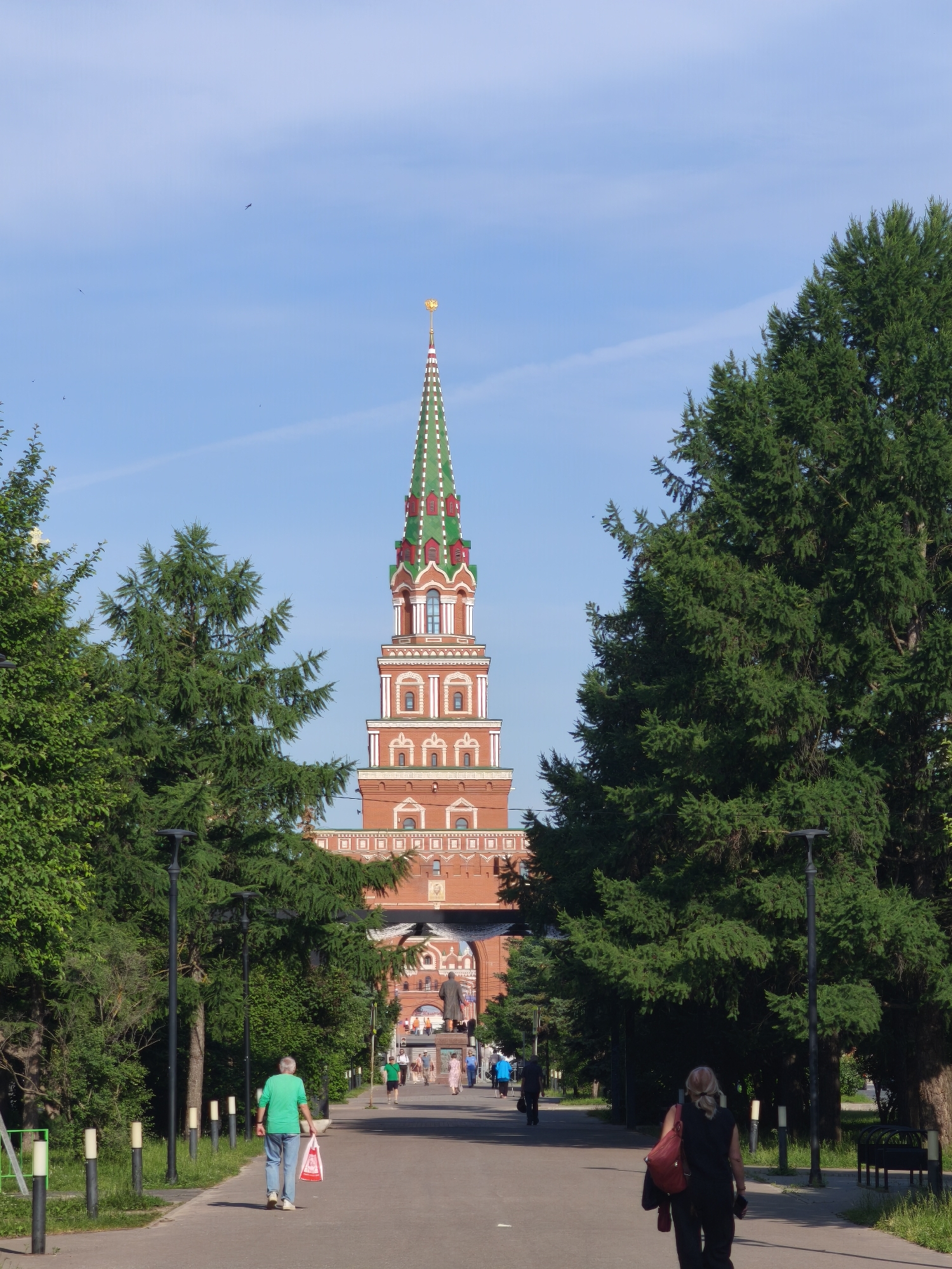 The image captures a frontal view of the Tsar Kokai Tower in Yoshkar-Ola, Russia, under a clear blue sky with a few wisps of clouds. The tower, constructed of red brick, features intricate white detailing, arched windows, and a green, multi-tiered spire topped with a golden element. The tower is framed by lush green trees on either side of a paved pathway.
In the foreground, a paved walkway leads towards the tower. On the left side of the walkway, a man in a green t-shirt and blue jeans walks away from the camera, carrying a red and white shopping bag. Several black lampposts with white lights line the left side of the path, interspersed with shorter, cylindrical white bollards. On the right side of the pathway, a woman with blonde hair pulled back, wearing a black sleeveless top and black pants, walks away from the camera, carrying a red backpack.
Further down the pathway, through an arched gateway at the base of the tower, a statue of a man is visible. Several other people are walking along the path in the distance, and more trees and greenery are visible beyond the archway. The overall scene depicts a sunny day in a well-maintained public space.