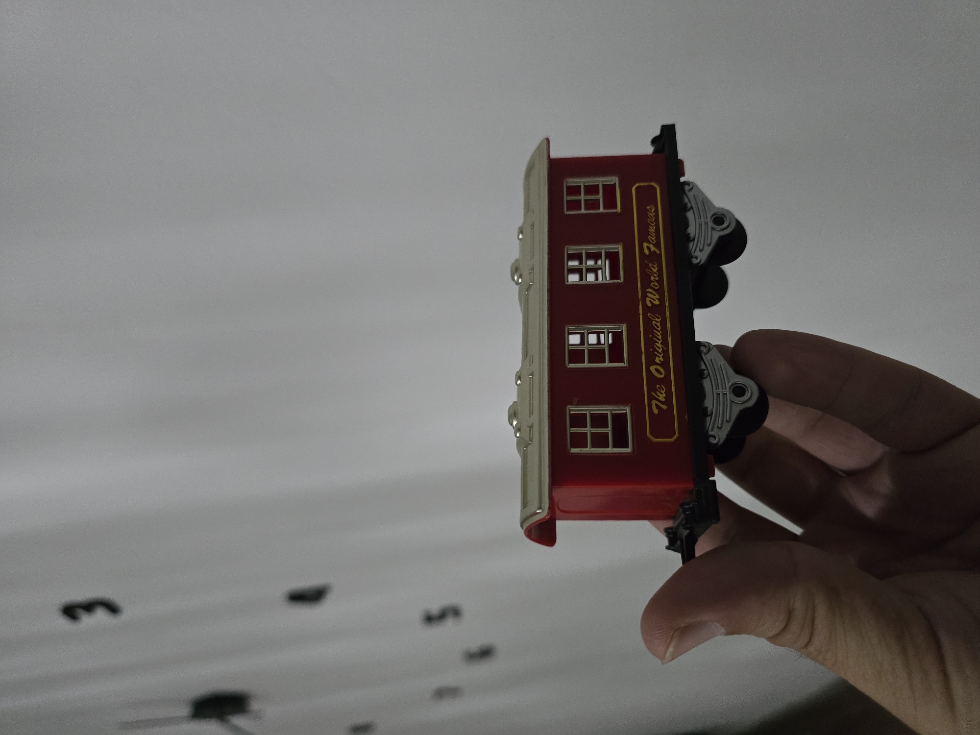 A hand is holding a red toy train car with a gold inscription that reads "The Original World Famous". The train car has three windows on its side and a silver roof with visible rivets. In the background, a wall is visible with faint black numbers that appear to be part of a clock face. The lighting suggests an indoor setting.