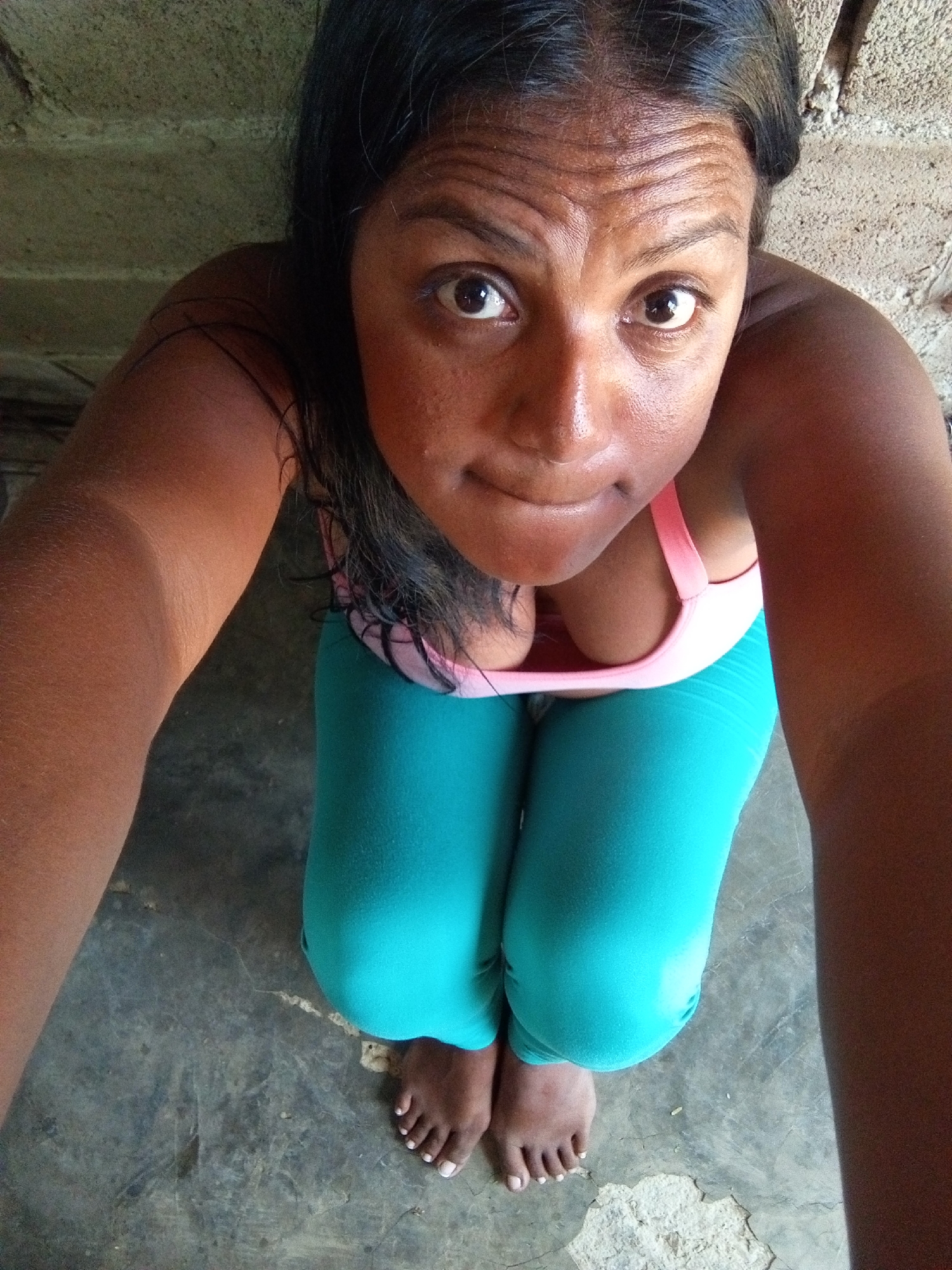 A high-angle, full shot depicts a woman kneeling on a grey, possibly concrete, floor. She is wearing a light pink sports bra and teal leggings. Her bare feet are visible at the bottom of the frame. She has dark hair and brown eyes and is looking up at the camera. The background is a mottled grey color, possibly a wall. Location is unknown.