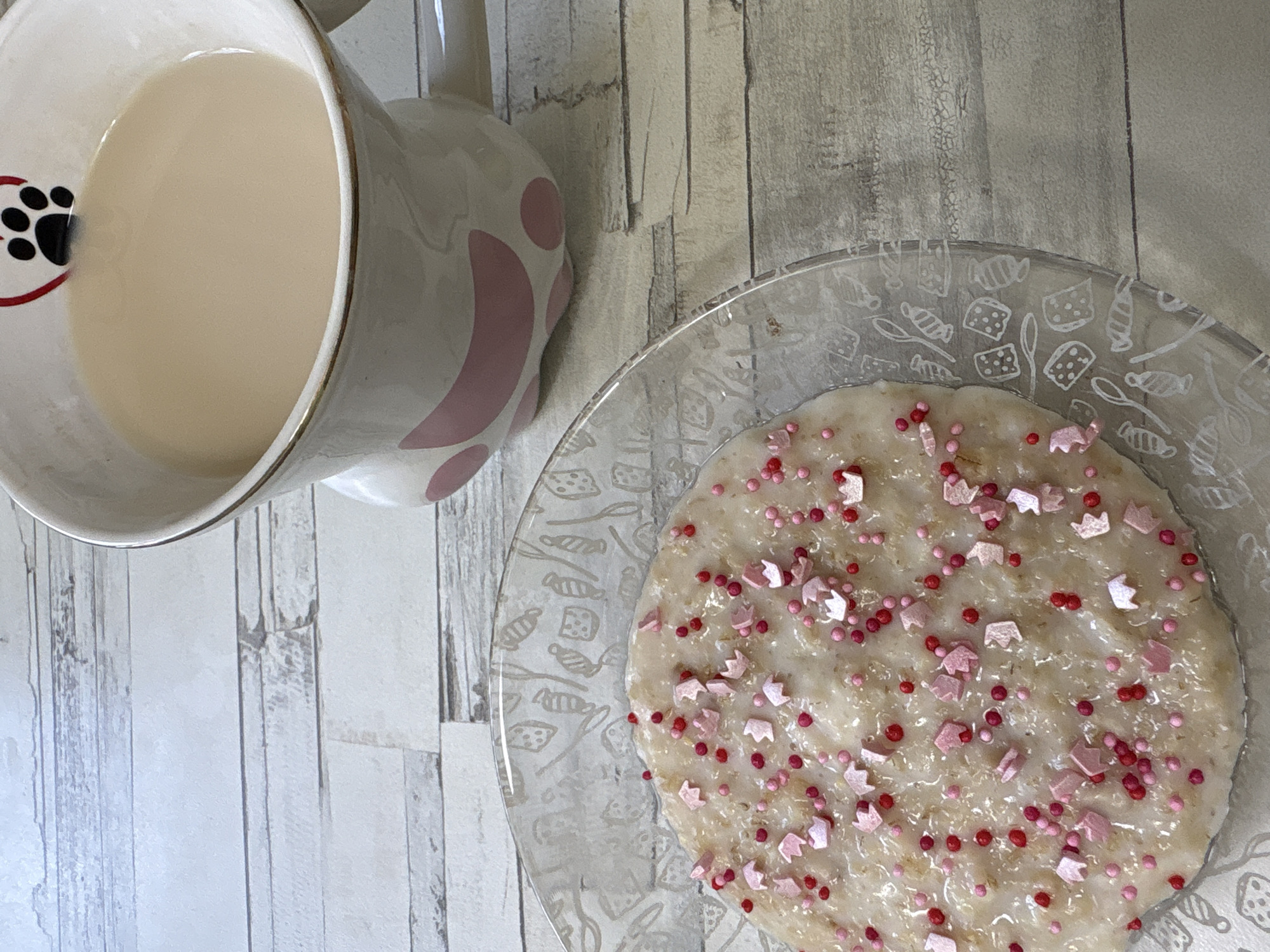 This top-down image captures a cozy breakfast or snack arrangement, likely set in a home in Chaikovsky, Russia. The scene unfolds on a light-colored wooden surface, characterized by distinct vertical planks and visible grain, suggesting a rustic or natural aesthetic.
In the upper left, a ceramic mug, predominantly white or cream, holds a light beige liquid, possibly milk, a latte, or tea with milk. The mug features charming details: a black paw print is depicted within a red heart on its interior rim, and subtle pink, abstract patterns adorn its exterior. A delicate gold line also highlights the mug's rim.
To the right, a clear glass bowl or shallow plate contains a creamy, light beige dish, which appears to be oatmeal, porridge, or rice pudding. This meal is whimsically garnished with an abundance of sprinkles. Two types are visible: numerous small, round sprinkles in shades of red and dark pink, and larger, distinct light pink sprinkles shaped like miniature crowns. The rim of the glass bowl itself is decorated with etched or embossed patterns featuring various confectionery shapes, including wrapped candies, squares that resemble cheese, and outlines of spoons and forks.
The image portrays a peaceful moment, hinting at a comfortable start to the day or a pleasant mid-day break. No individuals are present in the frame, and no visible text can be deciphered. The overall impression is one of simple indulgence and homeliness.