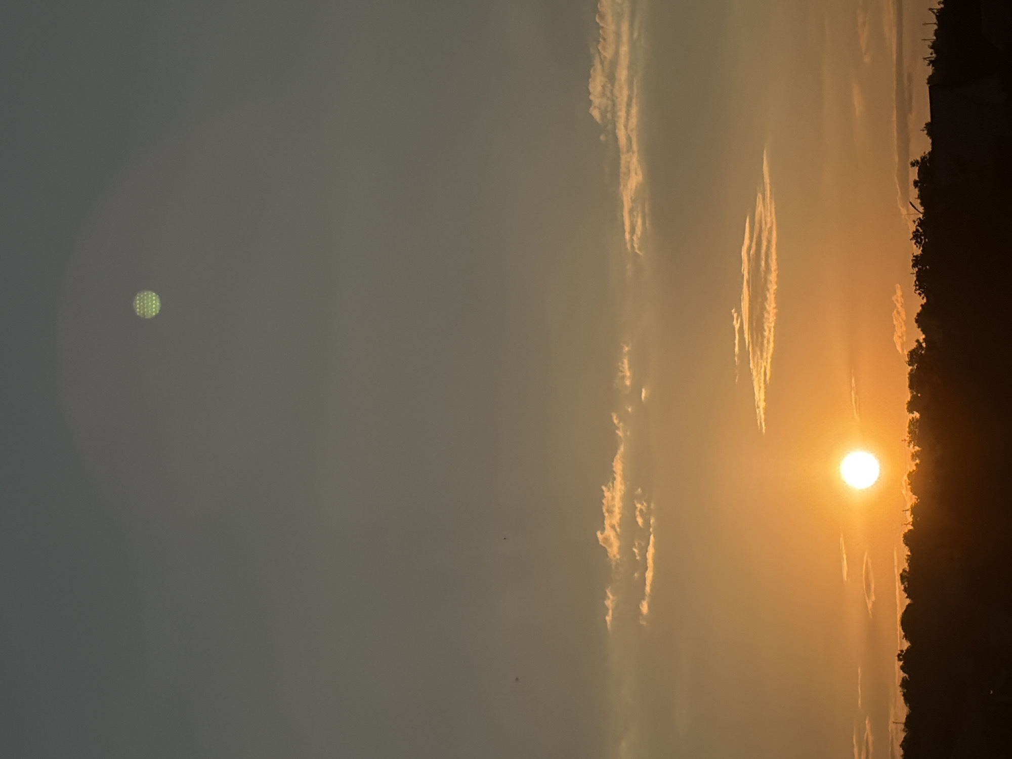 The image depicts a sunset over a silhouetted landscape in Oxentea, Moldova. The sky transitions from a pale orange near the horizon to a muted blue-gray at the top. The sun, a bright, glowing orb, is low in the sky, casting a warm orange light that illuminates a few scattered, wispy clouds. Below the sun, the horizon is a dark, indistinct silhouette of trees and possibly buildings, suggesting a rural or semi-urban setting. A distinct circular artifact, appearing as a grid of tiny green dots, is visible in the upper portion of the sky, likely a lens flare or digital artifact. The overall scene is tranquil, capturing the atmospheric conditions of twilight.