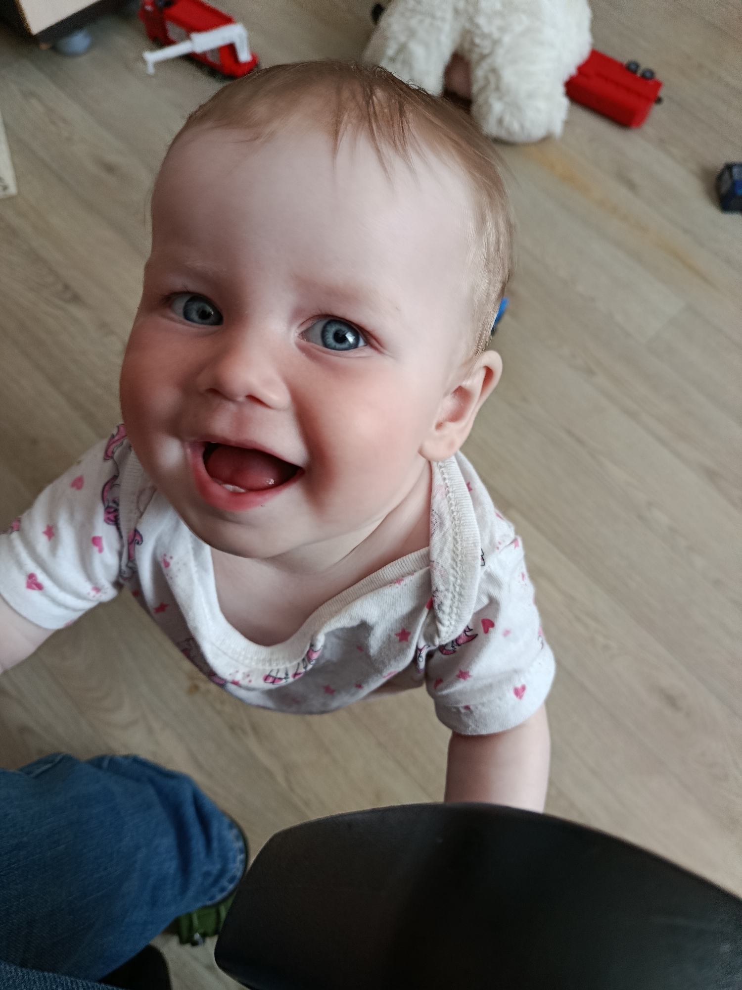 A baby with blue eyes and light brown hair is smiling at the camera. The baby is wearing a white onesie with pink hearts. The baby appears to be supported by a black object. In the background are children's toys, including a red toy truck and a white stuffed animal. The floor is a light brown wood laminate. The image location and city are not determinable.
