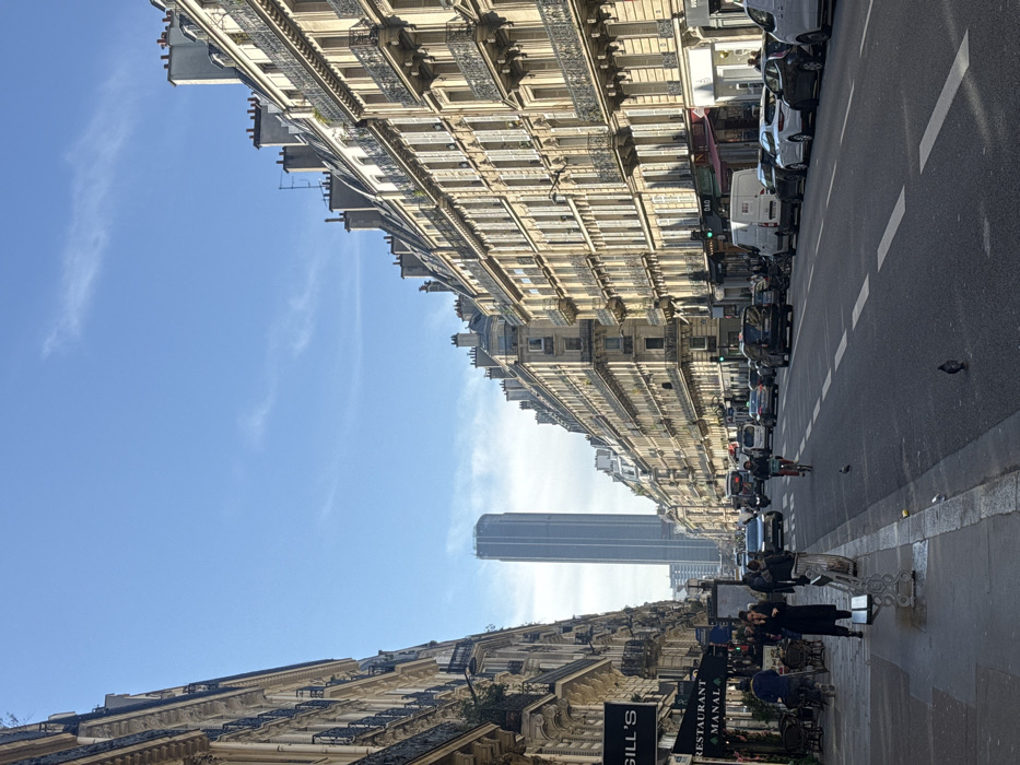 This image depicts a wide urban street, Rue de Rennes, in Paris, France, looking south directly towards the Tour Montparnasse.
The scene is set under a clear blue sky with sparse, wispy clouds, illuminated by natural sunlight that casts shadows. Both sides of the street are lined with multi-story, classic Parisian Haussmannian buildings featuring light-colored stone facades, numerous windows, and wrought-iron balconies. Rooftops are characterized by multiple chimneys. The ground floors of these buildings host various commercial establishments, including "GILL'S" and "RESTAURANT MANAL" visible on the left.
The street itself is a paved, multi-lane thoroughfare with white lane markings. Vehicular traffic includes numerous cars, a white delivery van, and a motorcycle. Some cars are parked along the right-hand curb. A cyclist in a light-colored top is visible in the middle lane of the road, proceeding away from the viewer.
On the left sidewalk, several pedestrians are present. One individual in a dark coat stands next to a white public bench, appearing to interact with a mobile device. Other pedestrians are visible further down the street, some near outdoor seating areas of restaurants. Pigeons are scattered on the sidewalk and the road surface.
The most prominent feature in the distance, framed by the converging buildings, is the Tour Montparnasse, a tall, slender, and grey cylindrical skyscraper, standing against the horizon. The overall scene depicts a busy daytime urban environment.