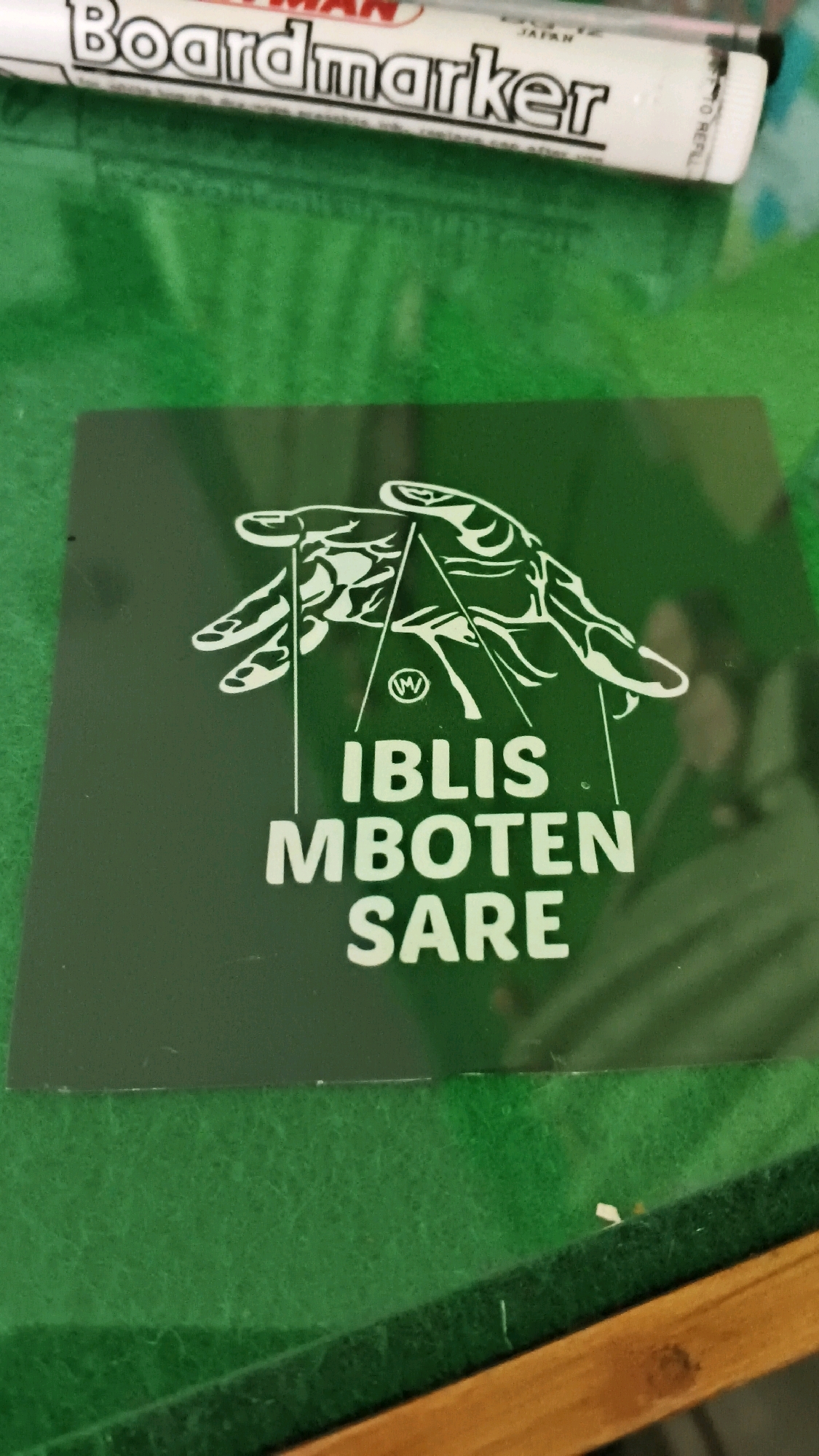 The image shows a dark green square sticker with white text and a white graphic design. The text reads "IBLIS MBOTEN SARE". Above the text is a graphic of two hands reaching upwards, with lines connecting them. In the center of the graphic, within a circle, is the letter "W". The sticker is placed on a green felt-like surface, possibly a table or a cue cloth. In the background, a white marker pen with the words "Boardmarker" and "JAPAN" is partially visible. The scene appears to be indoors, with some wood grain visible at the bottom edge of the frame. There is no information in the image to suggest a specific location in Karangmalang, Indonesia.