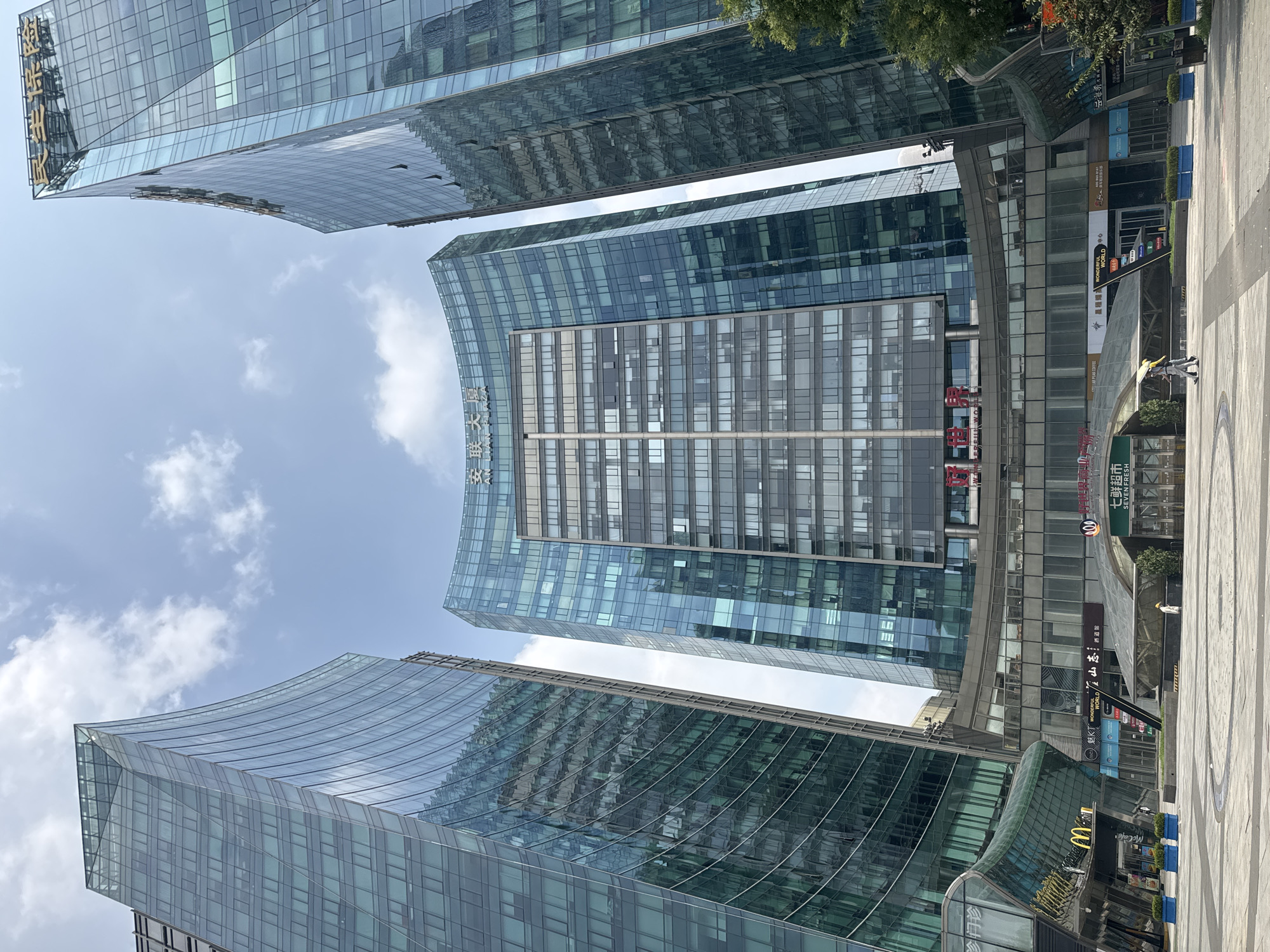 This is a wide-angle, low-angle shot of a modern urban complex in Chaoyang District, China, dominated by three towering glass buildings. The central building, a curved structure with a prominent golden inscription at its apex, is flanked by two taller, angular glass skyscrapers. The facades of all three buildings are reflective, mirroring the sky and clouds, and revealing the grid-like pattern of windows.
At the base of the buildings is a commercial area with storefronts, including a McDonald's with its distinctive golden arches logo. Other businesses are visible, with signage in Chinese. The ground level is paved with light-colored tiles, forming a plaza area. Two individuals are walking across the plaza: one person wearing a yellow hard hat and carrying an umbrella, and another person walking alongside them. Blue planters with green foliage are placed along the edge of the plaza. The sky above is a clear blue with scattered white clouds.