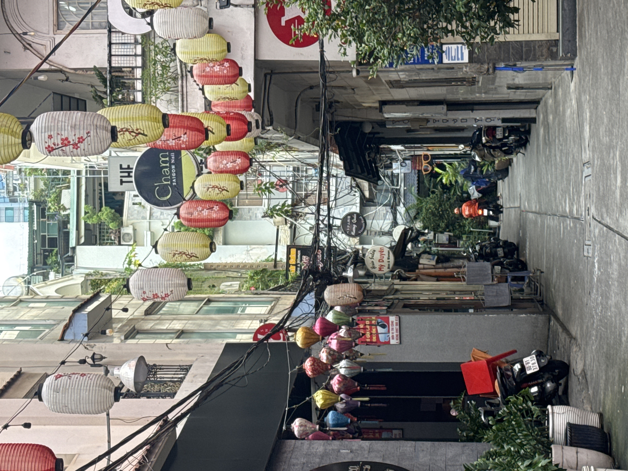 The image depicts a narrow street in the city of Thủ Đức, Vietnam, lined with buildings and adorned with numerous colorful paper lanterns. The street is paved with concrete and shows signs of wear. On the left side, a building with a light pink facade features balconies with intricate metal railings and is draped with electrical wires. Several white and red lanterns hang prominently, some with floral designs. Adjacent to this building is a darker facade, possibly a shop entrance, with potted plants in the foreground.
On the right side of the street, buildings present a more weathered appearance, with visible signs of age and various shop facades. A circular sign with "Cham SAIGON NAIL" is visible. Further down the street, a person wearing an orange jacket is seen riding a motorcycle away from the viewer, contributing to the sense of activity in the urban environment. Motorcycles are parked along the sides of the street, and more lanterns, in a variety of colors including yellow, pink, and blue, are suspended overhead, creating a festive and vibrant atmosphere. The architecture suggests a mix of traditional and modern elements, common in Vietnamese cities. Overhead, a complex network of electrical wires crisscrosses the sky, indicating the urban infrastructure. The lighting suggests it is daytime, with shadows cast on the ground.