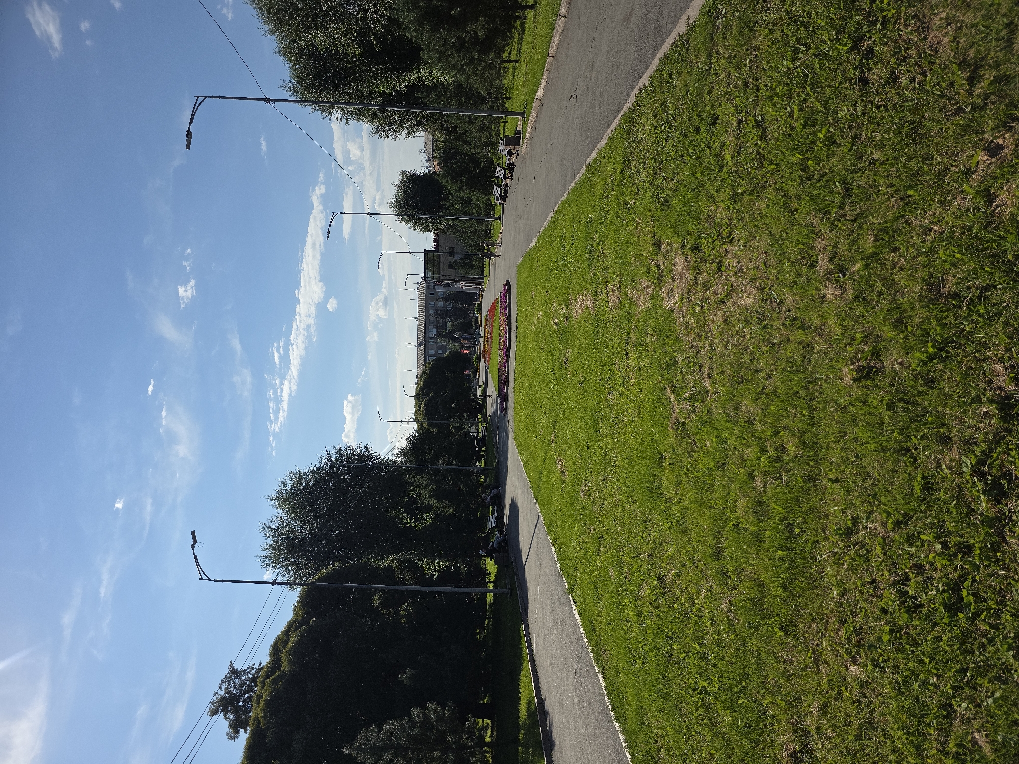 The image depicts a park scene in Chelyabinsk, Russia, likely during daytime. A wide, paved pathway curves to the right, bordered by a manicured green lawn on the left and more lawn and trees on the right. Several streetlights with angled lamps are visible along the pathway, indicating it's a public area. In the distance, a row of buildings and trees form the backdrop. A central flowerbed with red flowers is visible further down the path. A few individuals are seated on benches along the right side of the pathway, and others are walking in the distance. The sky is blue with scattered white clouds.