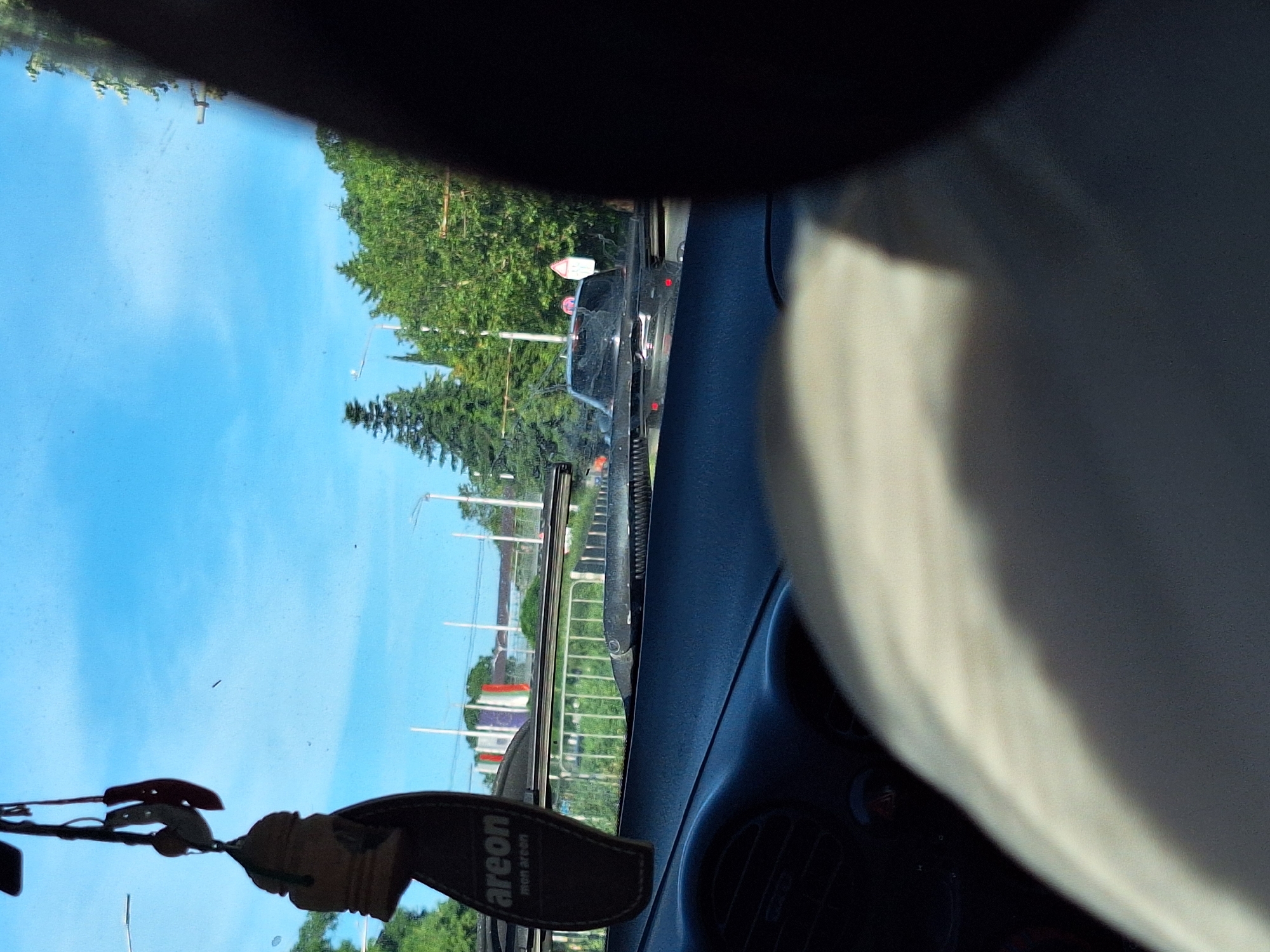 The image is a view from inside a car in Varna, Bulgaria. In the foreground, the dashboard and a portion of the driver's shoulder are visible. Hanging from the rearview mirror is a car air freshener. Through the windshield, traffic can be seen on a road lined with trees and streetlights. Several cars, including a dark-colored SUV, are on the road. In the background, buildings and flags are visible, suggesting an urban setting. The sky is clear and blue.