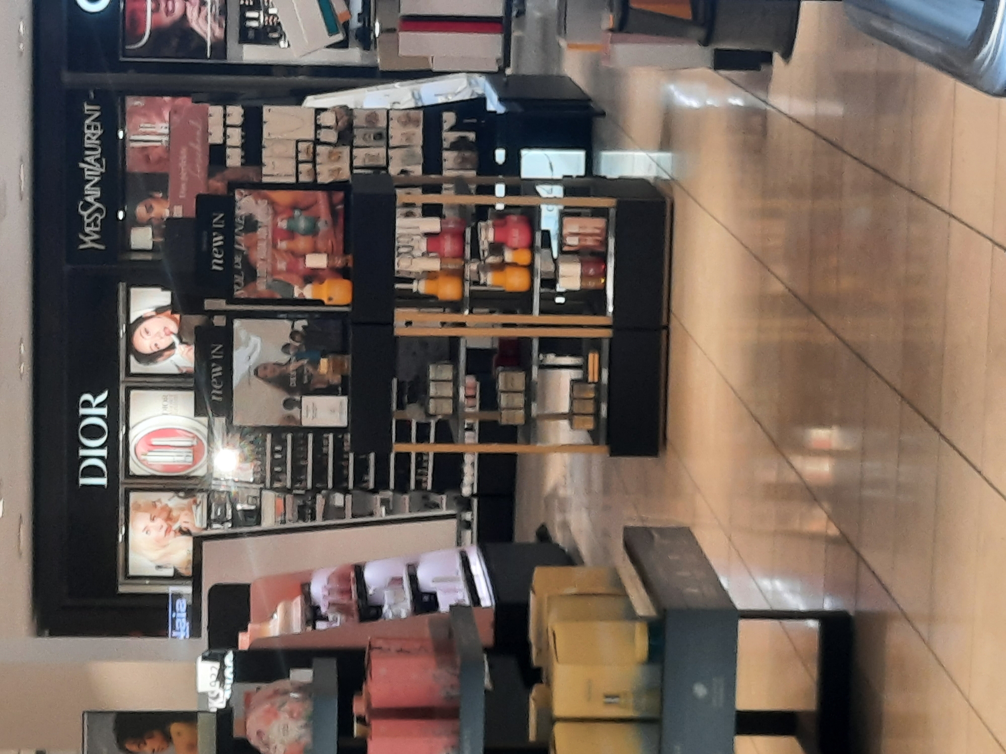 The image shows a retail store interior, likely a department store or a duty-free shop, featuring various cosmetic and fragrance brands. Prominent brands displayed include DIOR and YVES SAINT LAURENT. The store appears well-lit with a polished floor reflecting the overhead lighting. Several display shelves are visible, stocked with products like perfumes, makeup, and skincare items. The overall impression is one of a clean, modern retail environment. The provided context of Rosenheim, Germany, suggests this scene is located within a commercial establishment in that city.