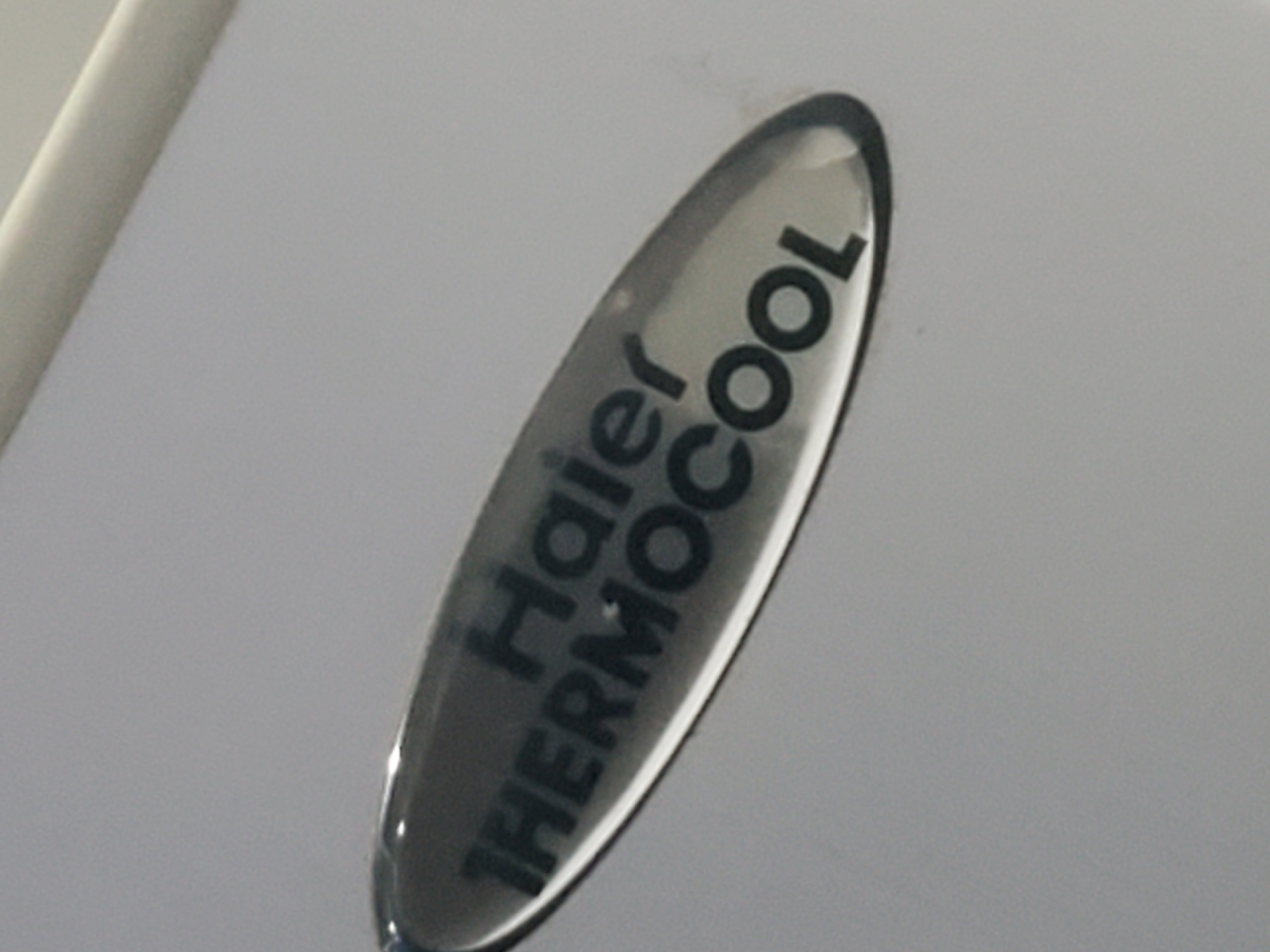 The image is a close-up, slightly angled shot of a metallic emblem on a light gray surface. The emblem is oval-shaped and has the text "Haier THERMOCOOL" inscribed on it in black, bold, sans-serif font. The emblem appears to be made of a reflective material, likely chrome or polished metal, with a black or dark gray insert for the text. The surface it's on is smooth and matte, with a subtle reflection visible on the left side of the emblem. The lighting suggests an indoor environment, with a soft, diffused light source. No people or other objects are visible in the frame. The location is unknown.