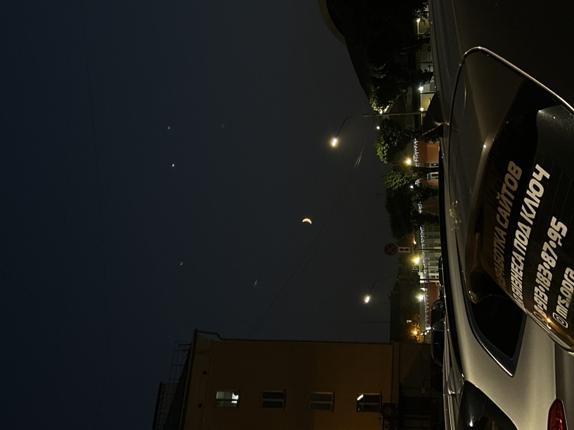 The image depicts a night scene in an urban environment. In the foreground, the rear of several parked cars is visible. One car has a white sticker on its rear windshield with text that appears to be in Russian, including a phone number and a social media handle. The middle ground features a street with streetlights, trees, and buildings. A crescent moon is visible in the dark sky. The background shows more buildings and the dark expanse of the night sky. The overall impression is of a quiet city street at night.