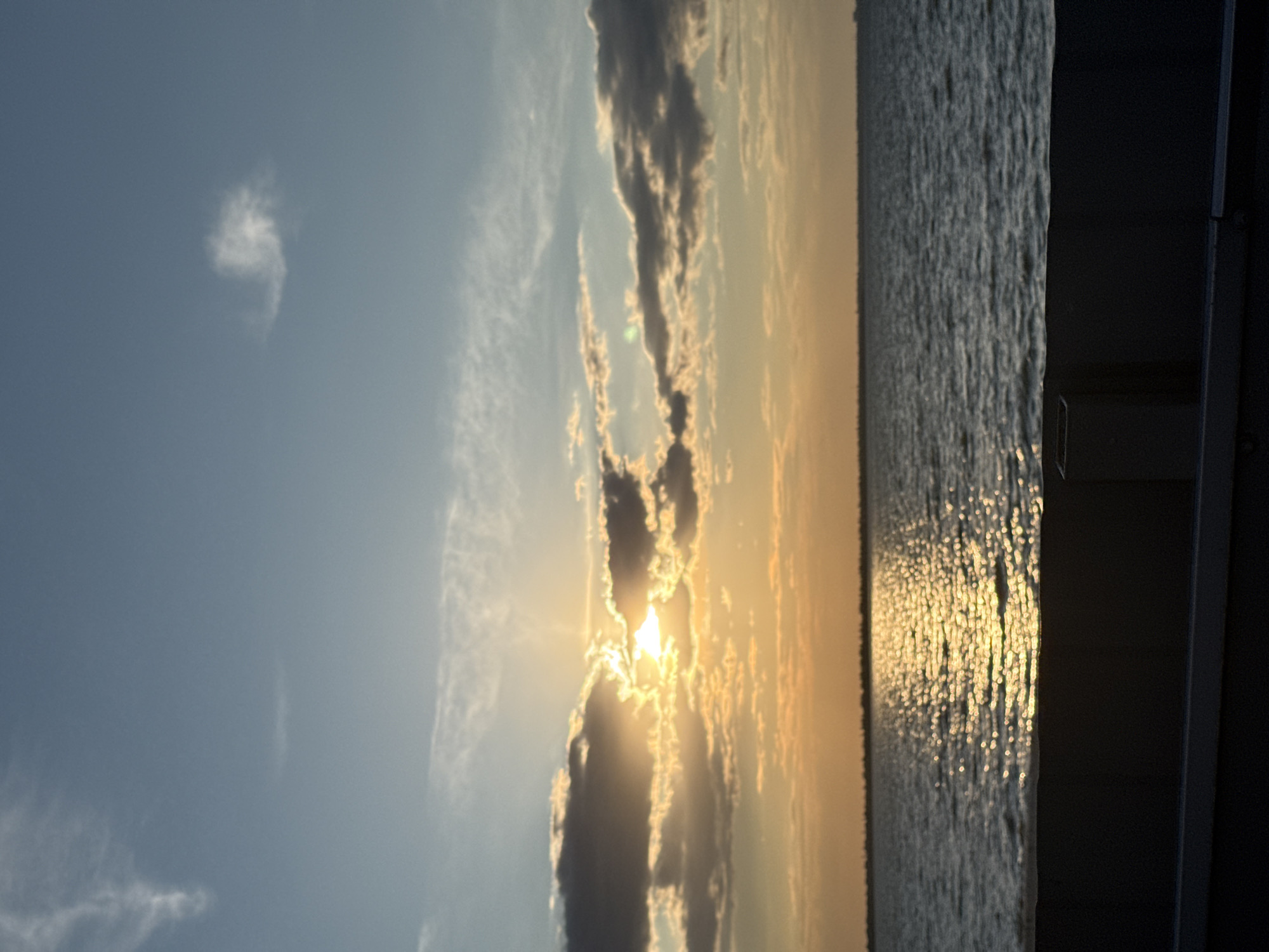 The image depicts a sunset over the Black Sea, likely taken from the promenade in Zatoka, Ukraine. The sun, partially obscured by clouds, casts a warm golden glow on the water, creating a shimmering reflection. The sky transitions from a pale blue at the top to hues of orange and yellow near the horizon, with scattered clouds. In the foreground, a dark, solid barrier runs horizontally across the bottom of the frame, with a small, dark trash receptacle visible on the right side of the barrier. The sea stretches to the horizon, where a faint, indistinct outline of buildings or land can be seen. The overall scene is tranquil and picturesque, characteristic of a coastal sunset.