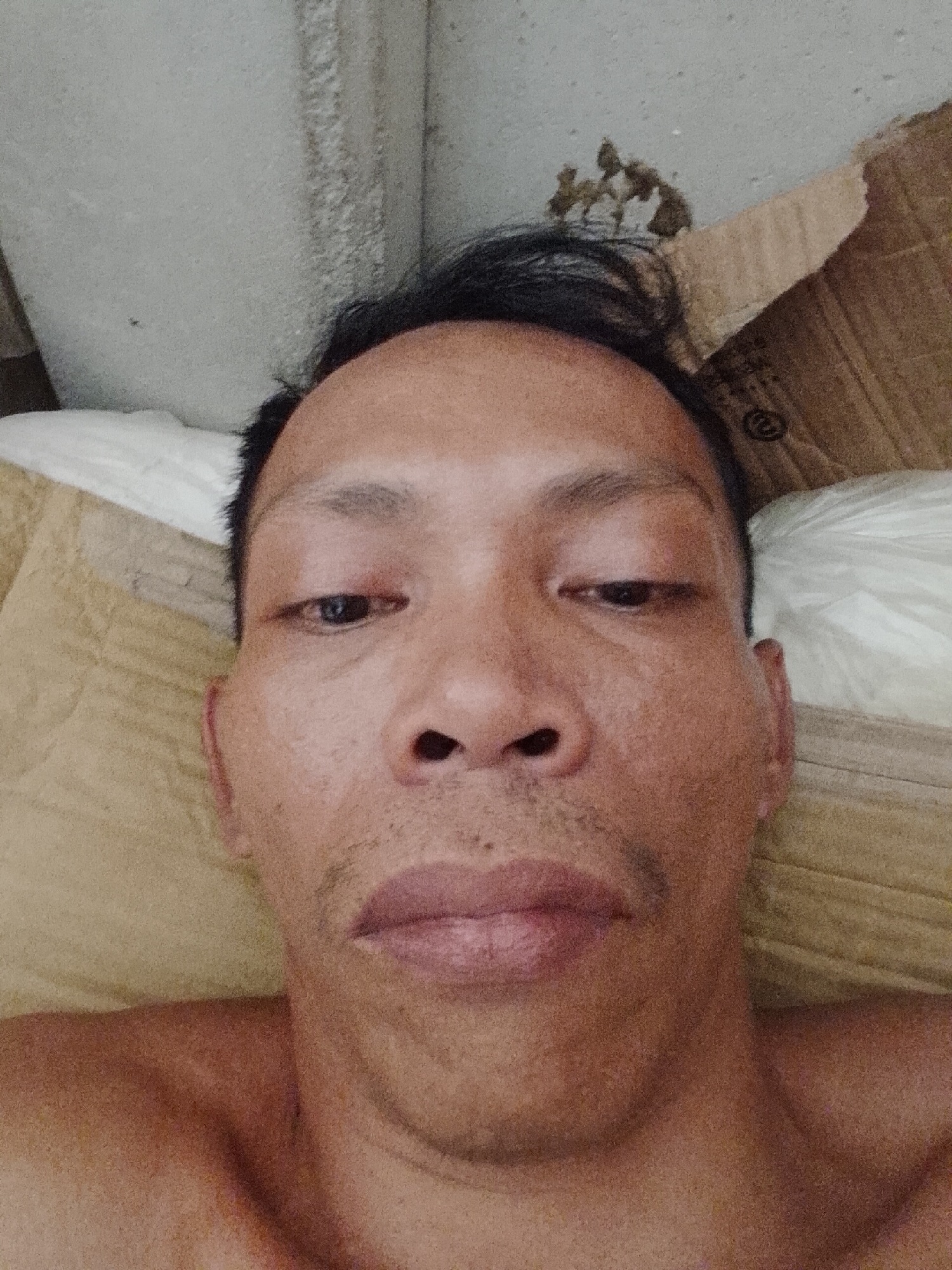 A close-up selfie of a middle-aged Asian man, lying down. He has dark hair with some sticking up, dark eyes, a wide nose, and a thin mustache. His skin appears oily. He is shirtless. He is lying on a tan-colored surface that appears to be a cushion or bedding. Behind him is a white pillow and a piece of cardboard. The background includes a portion of a white wall. There is no verifiable location information within the image. Therefore, no information is available to verify the location within the city of Bukit Ketok, Indonesia.