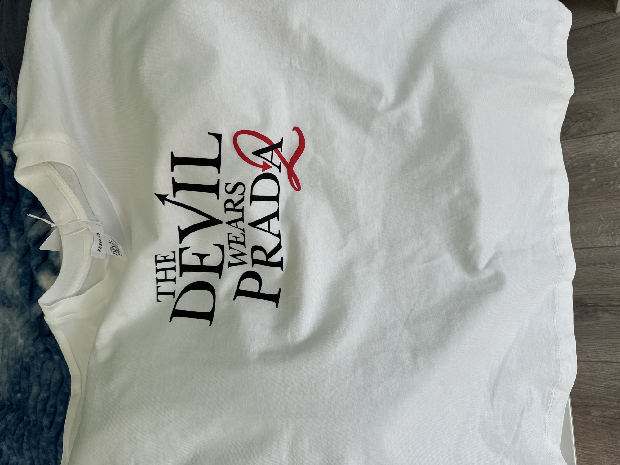 The image shows a white T-shirt with the text "THE DEVIL WEARS PRADA 2" printed on it, with the number "2" stylized in red. The shirt appears to be lying flat on a surface, with a partial view of a blue patterned fabric visible on the left side and wood floor on the right side. A tag is attached to the shirt's collar. There is no information to suggest the location is in Sofia, Bulgaria.