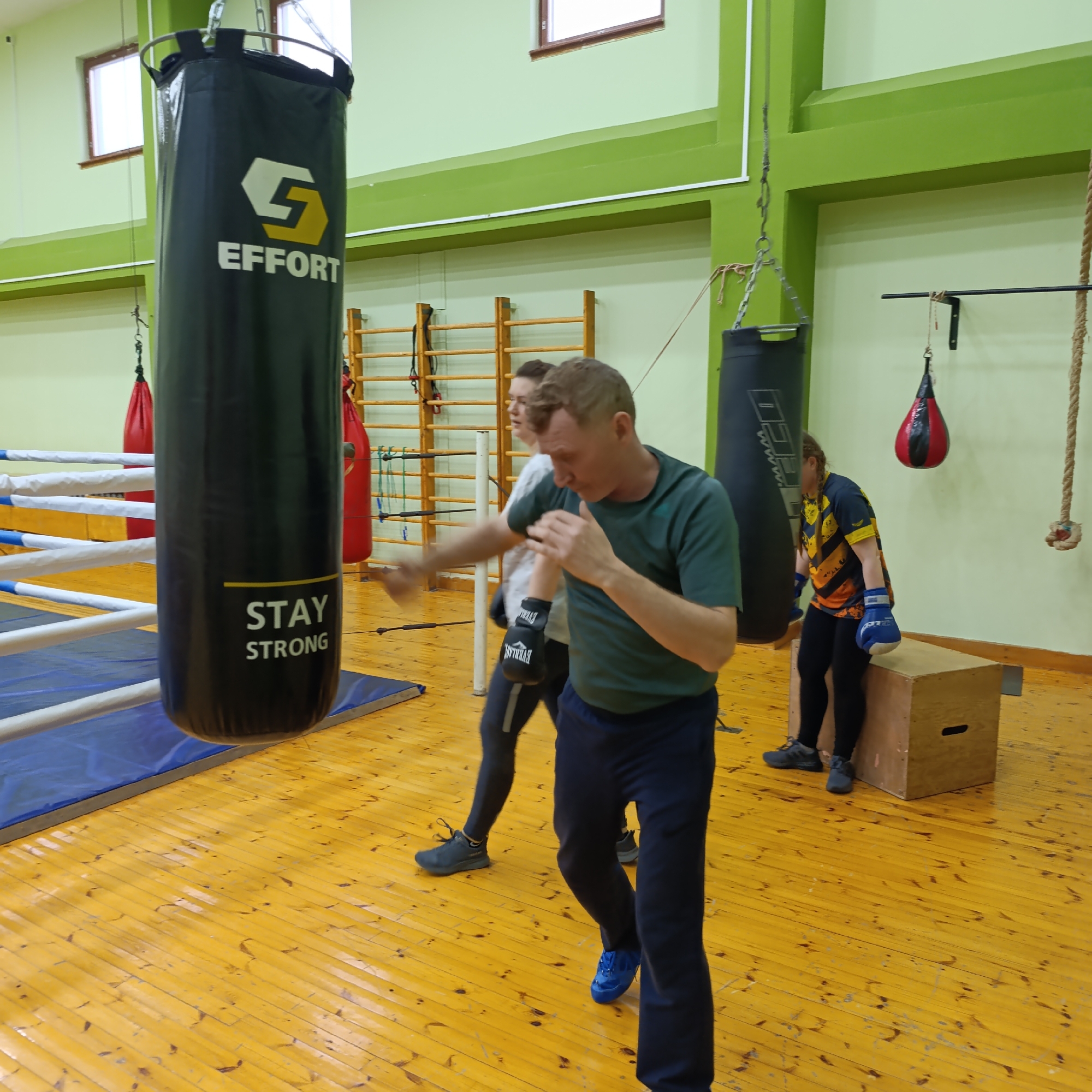 An active boxing training session is underway in an indoor gym in Markovskii, Russia. The room features light green walls with a darker green beam, a glossy wooden floor, and natural light entering through windows high on the walls.
In the foreground, a man with light brown hair, wearing a dark green t-shirt and dark blue pants, is punching a large black heavy bag with his right hand, which is protected by a black boxing glove. His left hand is held up in a guard position, and his expression is focused. The heavy bag is branded with a stylized 'G' logo, the word "EFFORT," and "STAY STRONG." Visible on his glove is the brand "EVERLAST."
Behind him, another individual with dark hair, wearing a white t-shirt and dark leggings, stands in a boxing stance, also with a black boxing glove on their right hand, appearing ready to train. To the far left, a section of a boxing ring with blue and white ropes and blue matting is visible.
In the background right, a third person, with braided hair and wearing a black and yellow/orange patterned top, black leggings, and blue boxing gloves, is seated on a wooden plyometric box. They are observing the activity.
Additional gym equipment includes a smaller black heavy bag, two red teardrop-shaped punching bags hanging from the ceiling, wooden wall bars, and a thick climbing rope, all distributed around the training space. The overall atmosphere suggests a dedicated and active training environment.