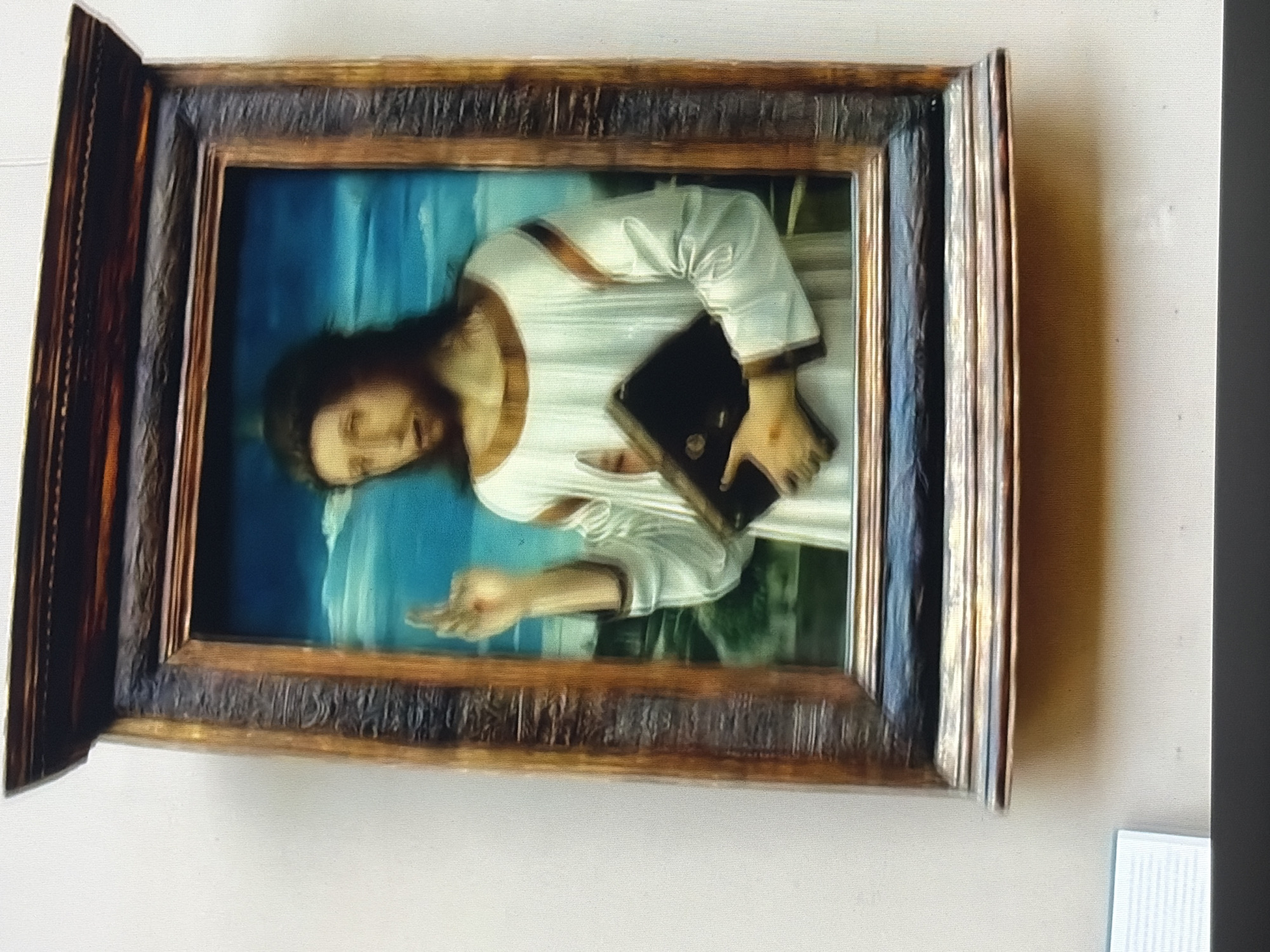 A painting of Jesus Christ is displayed in a museum. Jesus is depicted as a young man with long, dark hair and a beard. He is wearing a white robe with a brown trim around the neckline and sleeves. He is holding an open book in his left hand and has his right hand raised in a blessing gesture. The background of the painting is a bright blue sky with white clouds and a green landscape. The painting is housed in an ornate, dark wood frame with gold accents. The painting is hanging on a light-colored wall, and there is a shadow cast on the wall below it.