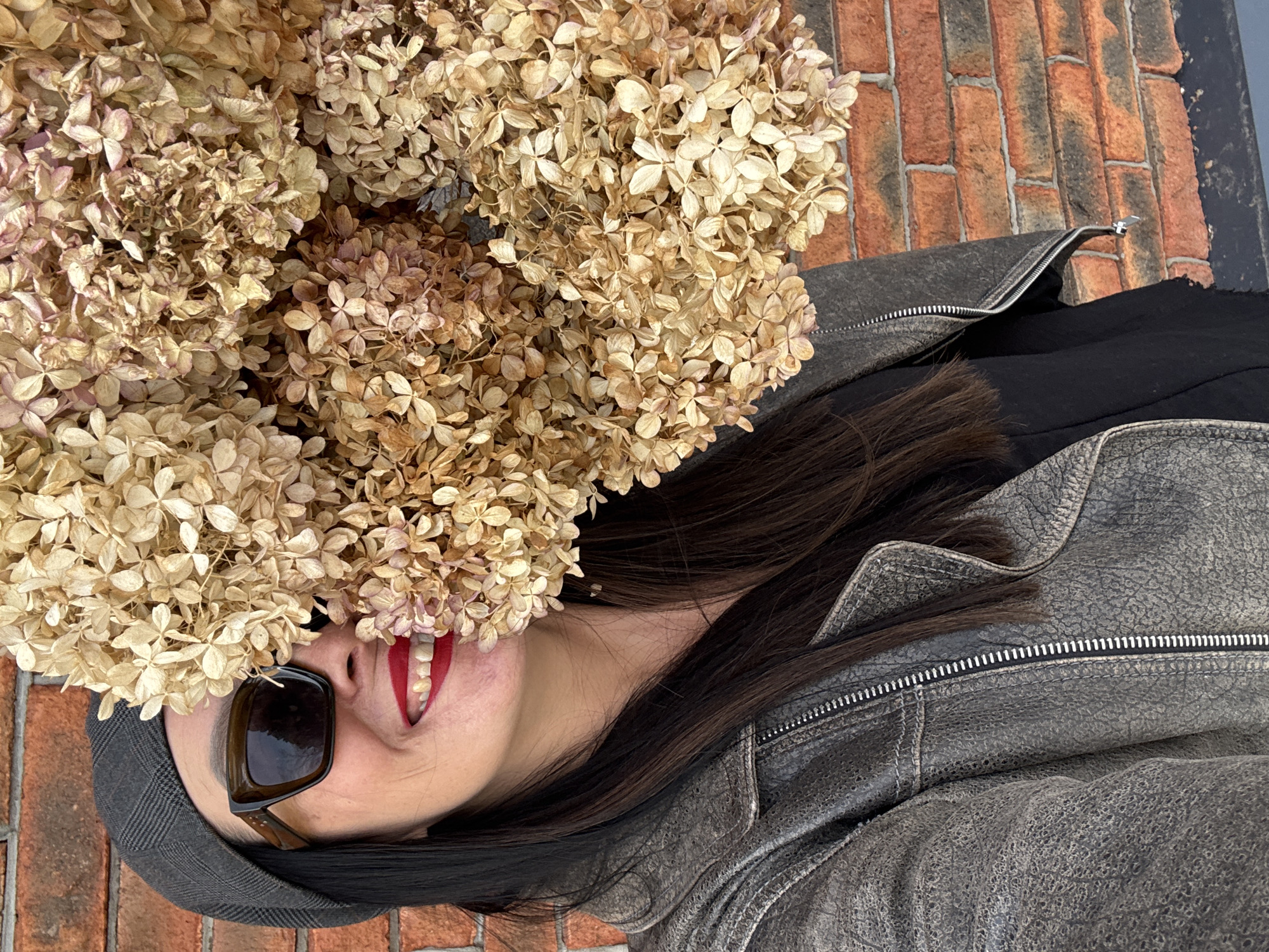A person wearing sunglasses and a cap is lying on a brick surface, partially covered by dried hydrangea flowers. The person has dark hair and is wearing a gray, weathered leather jacket over a black garment. Only part of their face, including their mouth with red lipstick, is visible among the flowers. The location appears to be outdoors. There is no specific information in the image to confirm this is the city of Chaikovsky, Russia.