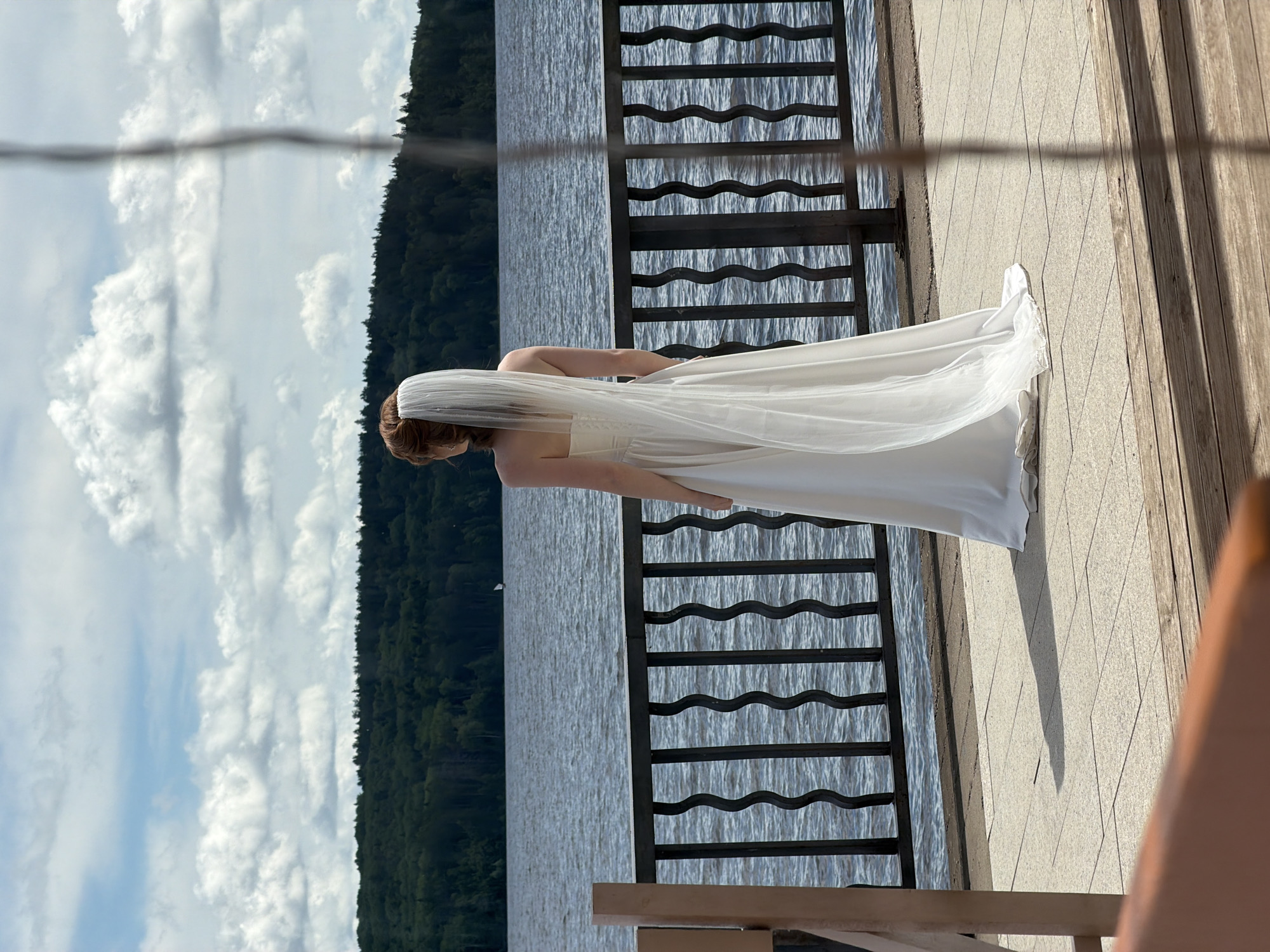 The image depicts a woman in a white wedding dress and veil, standing on a paved walkway next to a dark railing. Beyond the railing is a body of water with gentle waves, and in the distance, a tree-covered shoreline. The sky above is blue with scattered white clouds. The scene appears to be an outdoor location, possibly a waterfront promenade or a park. There are no visible landmarks in the image that would specifically place it in Chaikovsky, Russia.