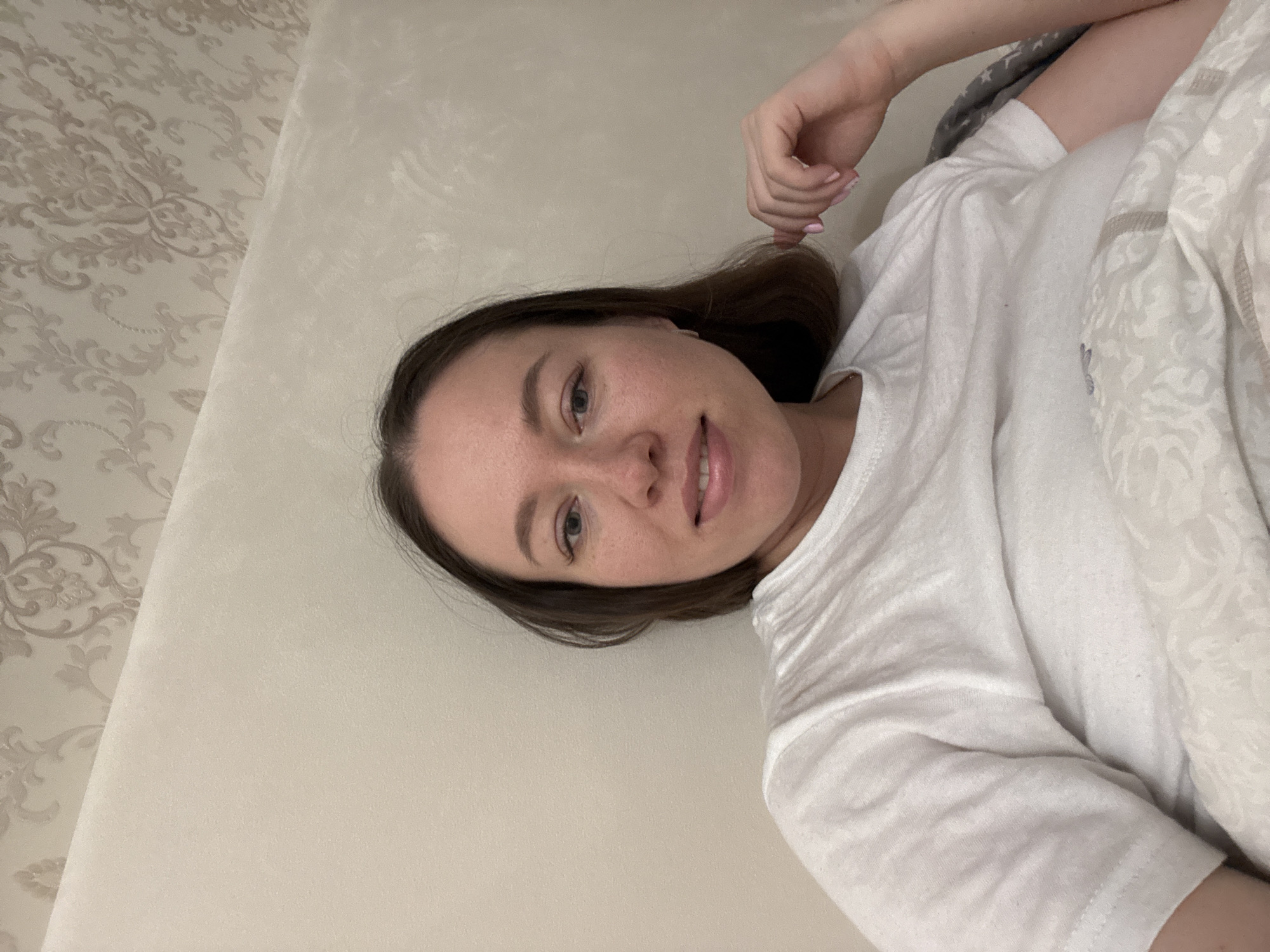 A woman lies on a beige-colored surface. She is wearing a white T-shirt. She has brown hair and light skin. Her right hand is raised near her head. She appears to be in a room with wallpaper. The location is potentially in Ферма, Russia.