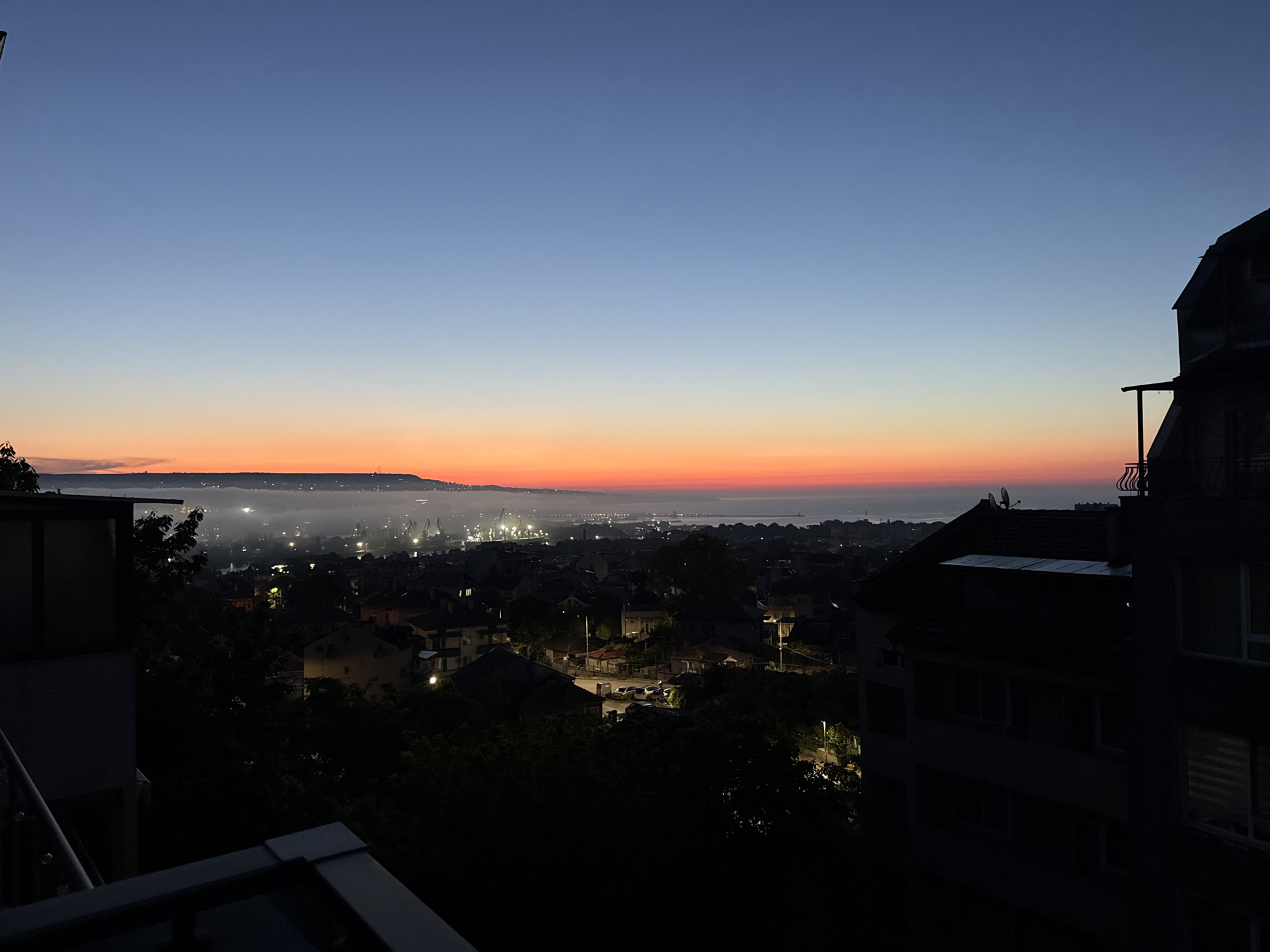 A panoramic view captures a city skyline at dusk. The sky transitions from a deep blue at the zenith to a vibrant orange and red hue along the horizon, indicating either sunrise or sunset. A layer of mist or fog blankets the lower portion of the city, obscuring some of the details and creating a soft glow from the streetlights and industrial illumination in the distance. Silhouetted buildings and trees dominate the foreground, suggesting an elevated viewpoint, possibly from a balcony or higher floor of a building. The architecture appears to be a mix of residential and possibly industrial structures, with cranes visible in the harbor area. The lighting is dim, with artificial lights from the city providing scattered points of illumination against the darkening landscape.