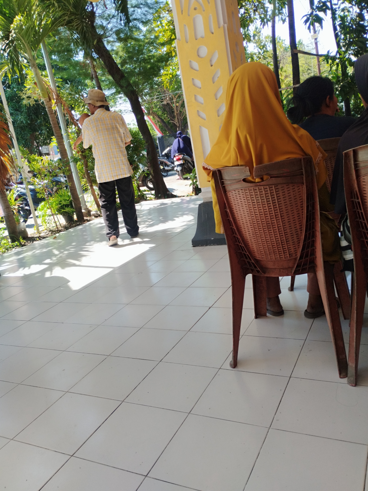 The image depicts an outdoor seating area, likely a cafe or restaurant, with a tiled floor and a yellow decorative wall. Several individuals are present. In the foreground, a person wearing a bright yellow headscarf sits in a brown plastic chair, facing away from the camera. To their right, another person wearing a black headscarf is partially visible. Further to the left, a man in a checkered shirt and hat walks away from the camera. Several motorcycles are parked in the background, suggesting a public space in a city like Surabaya, Indonesia. The scene is bathed in sunlight, casting shadows on the tiled floor, and surrounded by lush greenery.