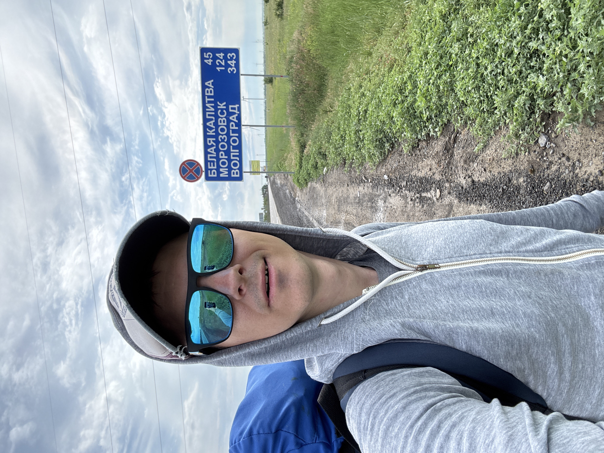 A selfie is taken by a person wearing a grey hooded sweatshirt, a baseball cap, and sunglasses. The person is carrying a large blue bag on their back and a backpack. The background shows a road, a grassy field with green vegetation, and a signpost with text in Russian and numbers. The sign indicates distances to "BELAYA KALITVA" (45 km), "MOROZOVSK" (124 km), and "VOLGOGRAD" (343 km). There is also a red and white circular traffic sign with a blue cross in the center. The sky is cloudy. The image appears to be taken outdoors in a rural or roadside setting, possibly on the outskirts of Verkhnegoveinyi, Russia, based on the Russian language on the signpost.