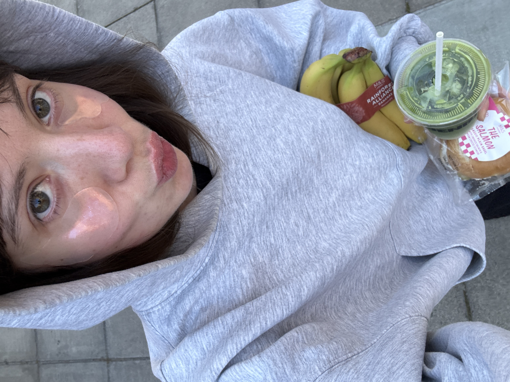 The image shows a selfie taken by a young woman with brown hair and green eyes. She has pink under-eye patches on her face. She is wearing a gray hoodie. Inside the hood of the hoodie, she is holding a bunch of bananas with a "M&S RAINFOREST ALLIANCE BANANAS" sticker on it. Next to the bananas, there is a plastic cup with a green beverage and a white straw. There is also a "THE SALMON" bagel sandwich, with "M&S" branding, visible. The background shows a gray, possibly concrete, ground, suggesting an outdoor location, possibly in the city of London, United Kingdom based on the M&S branding.
