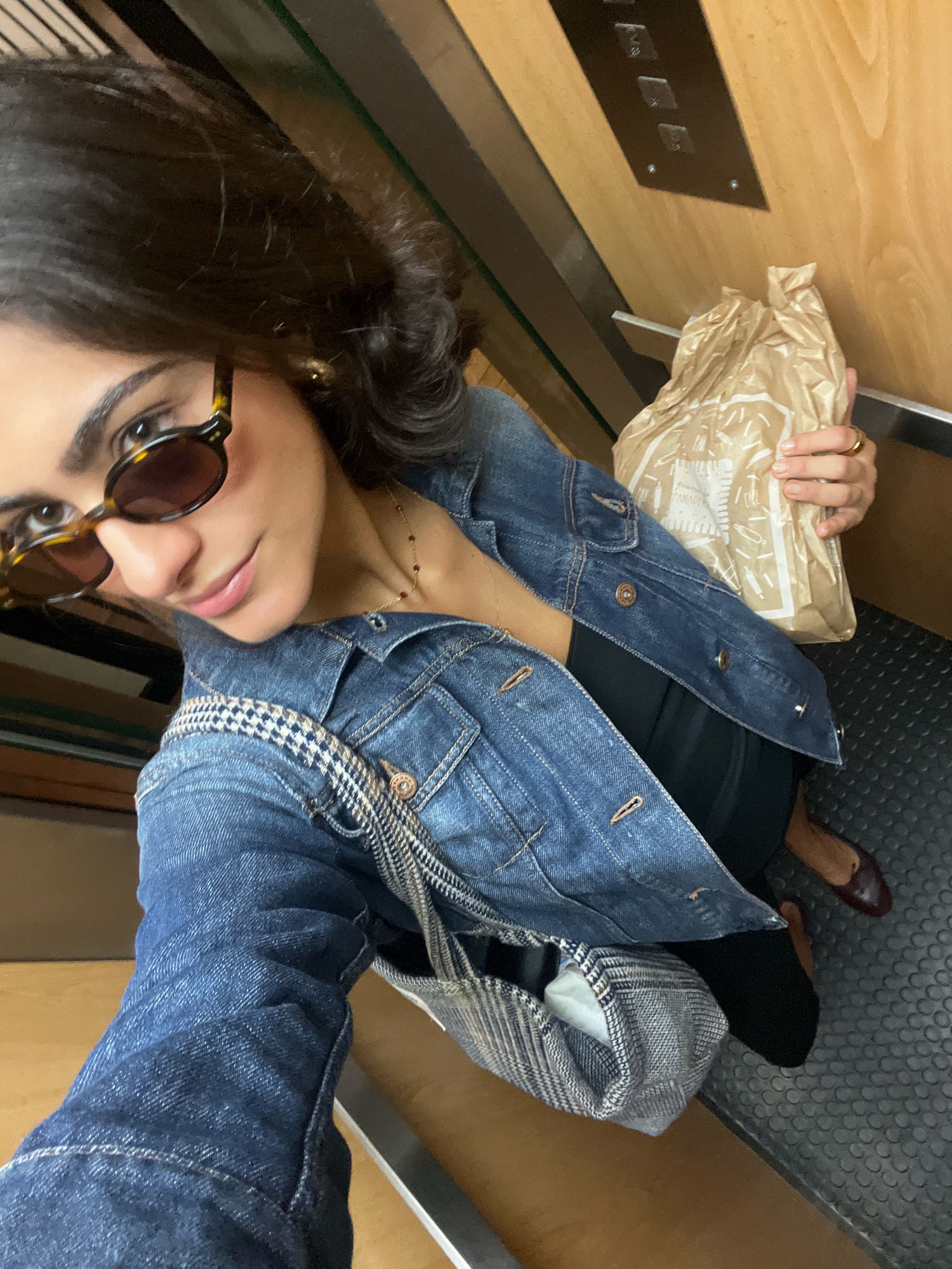A woman is taking a selfie in an elevator. She is wearing sunglasses, a denim jacket, and carrying a brown paper bag in her right hand. The elevator has wooden paneling and metal buttons on the wall. The floor is a gray, textured material. The woman is looking at the camera and smiling. The scene appears to be indoors, possibly in a residential building or hotel. There is no indication that this location is in the city of Madrid, Spain.