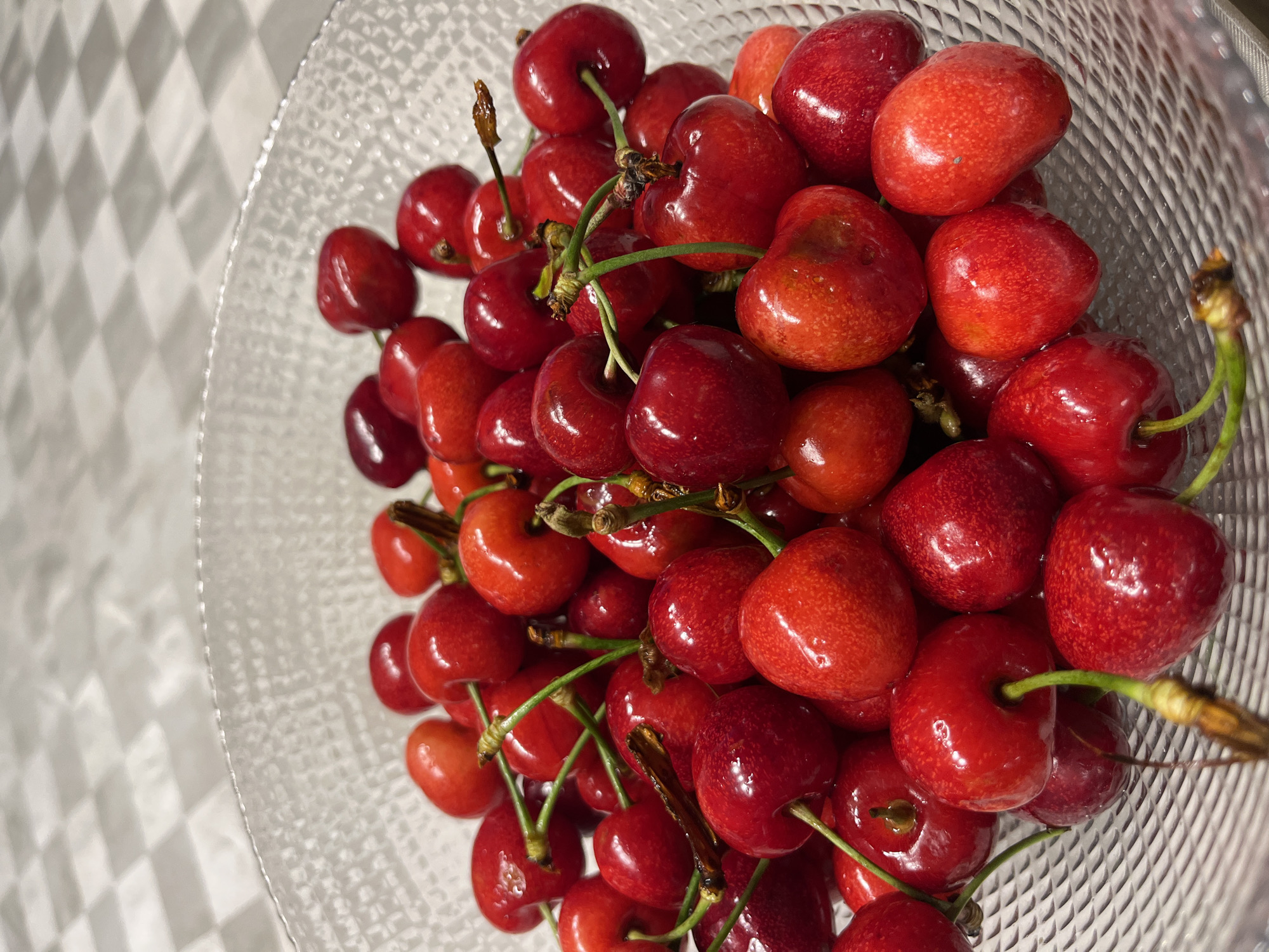 A close-up, eye-level shot captures a clear glass bowl filled with ripe, red cherries. The cherries are a deep, glossy red, with some exhibiting slight variations in color, including lighter red or pinkish hues. They are packed tightly together, with many still attached to their thin, green stems. The bowl is textured with a crisscross pattern, and it sits on a surface with a checkered pattern, possibly a tablecloth. The background is softly blurred, suggesting a shallow depth of field. The lighting is even, highlighting the smooth, lustrous surfaces of the fruit.