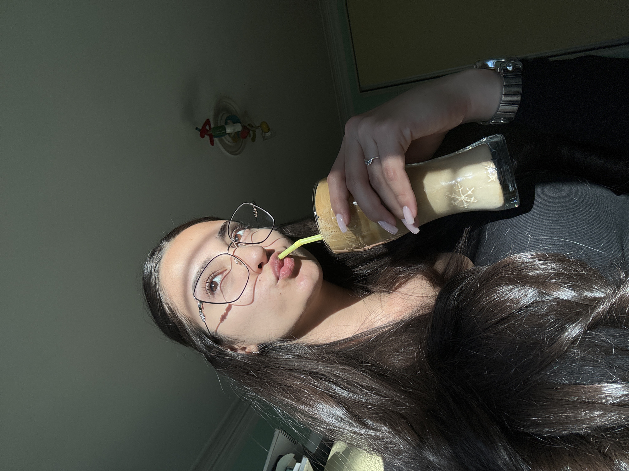 A young woman with long, dark hair is drinking from a clear glass with a straw. She is wearing glasses and a black shirt. Her nails are long and painted pink. She has a silver ring on her finger and a silver watch on her wrist. The background is blurry, but it appears to be a room with a light-colored wall. A decorative light fixture with colorful figures hangs on the wall. It is impossible to determine if the photo was taken in Dupnitsa, Bulgaria.
