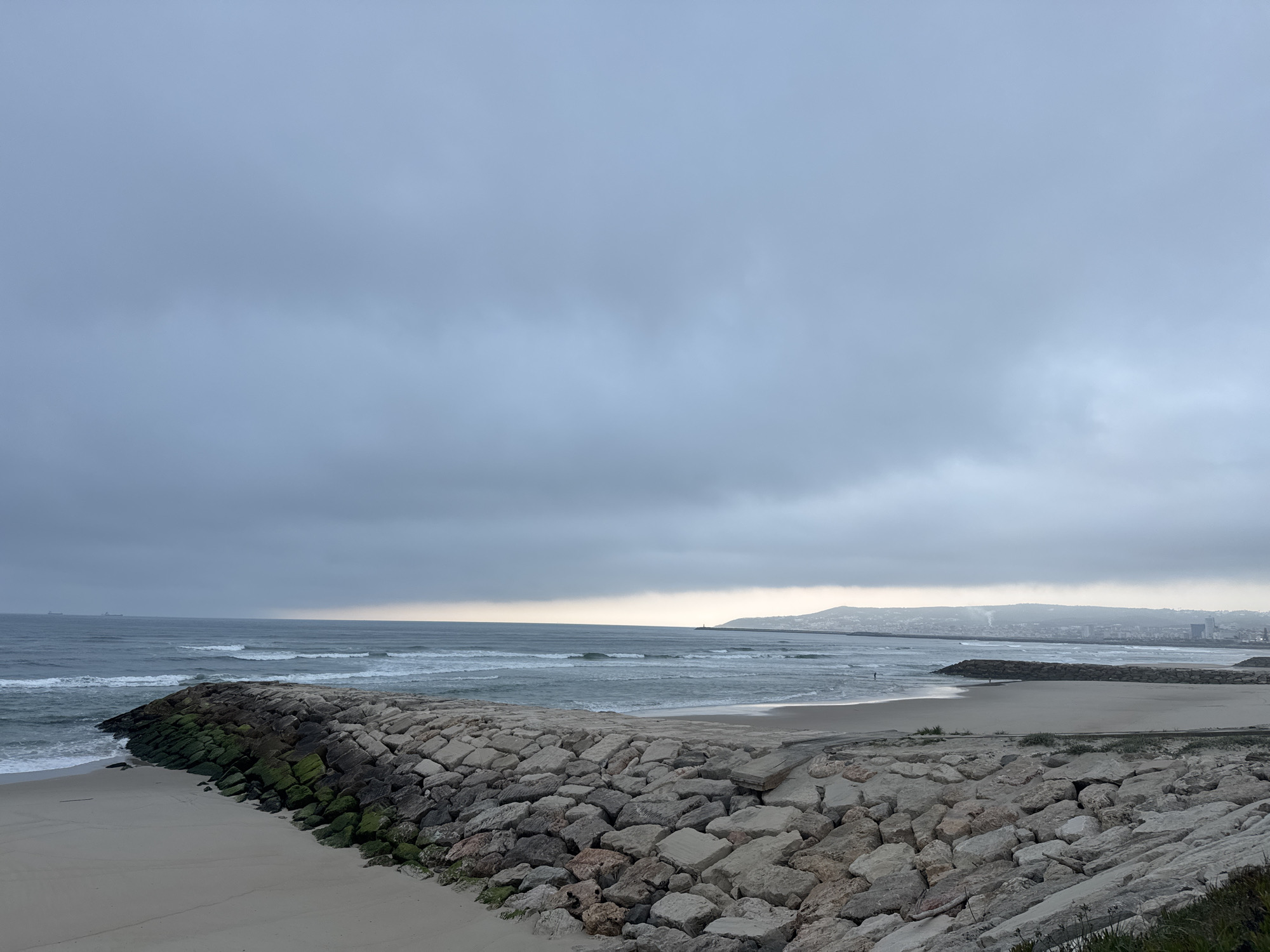 This media file captures a wide coastal view of Figueira da Foz, Portugal, on an overcast day. The scene is dominated by a long, curving rocky breakwater that extends into the ocean from a sandy beach. Many of the rocks, particularly those closer to the water, are covered in green moss or algae.
The ocean, with gentle waves breaking, stretches into the distance, and several large cargo or tanker ships are visible on the horizon. The sky is heavily clouded and grey, casting a diffused, muted light over the landscape. A slightly lighter band of sky is visible along the horizon, hinting at the sun's position either in the early morning or late afternoon.
To the right of the main breakwater, the sandy beach continues into the distance, where a lone, small figure can be seen standing near the water's edge. In the far background, a hilly coastline rises, featuring a distant cityscape or town dotted with buildings and a prominent lighthouse on a headland. No visible text is present in the image. The overall atmosphere is tranquil and calm, characteristic of a serene day at the coast.