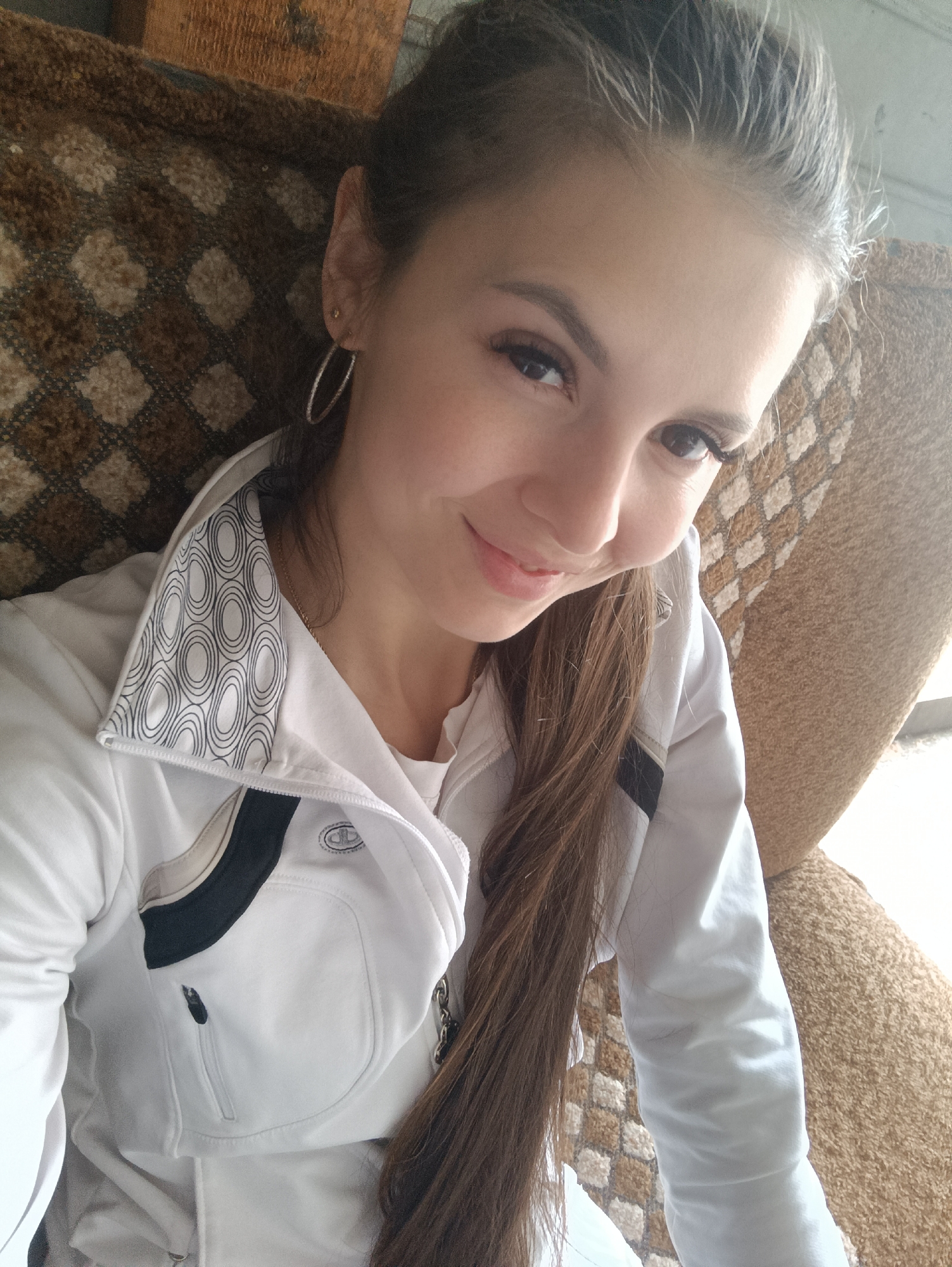A young woman is sitting on a patterned armchair in an indoor setting, possibly a home. She is fair-skinned with light brown hair pulled back into a ponytail, with some strands hanging down. She is wearing a white jacket with a patterned collar and black accents over a white t-shirt. She has hoop earrings and is looking at the camera with a slight smile. The location is Unknown, Unknown.