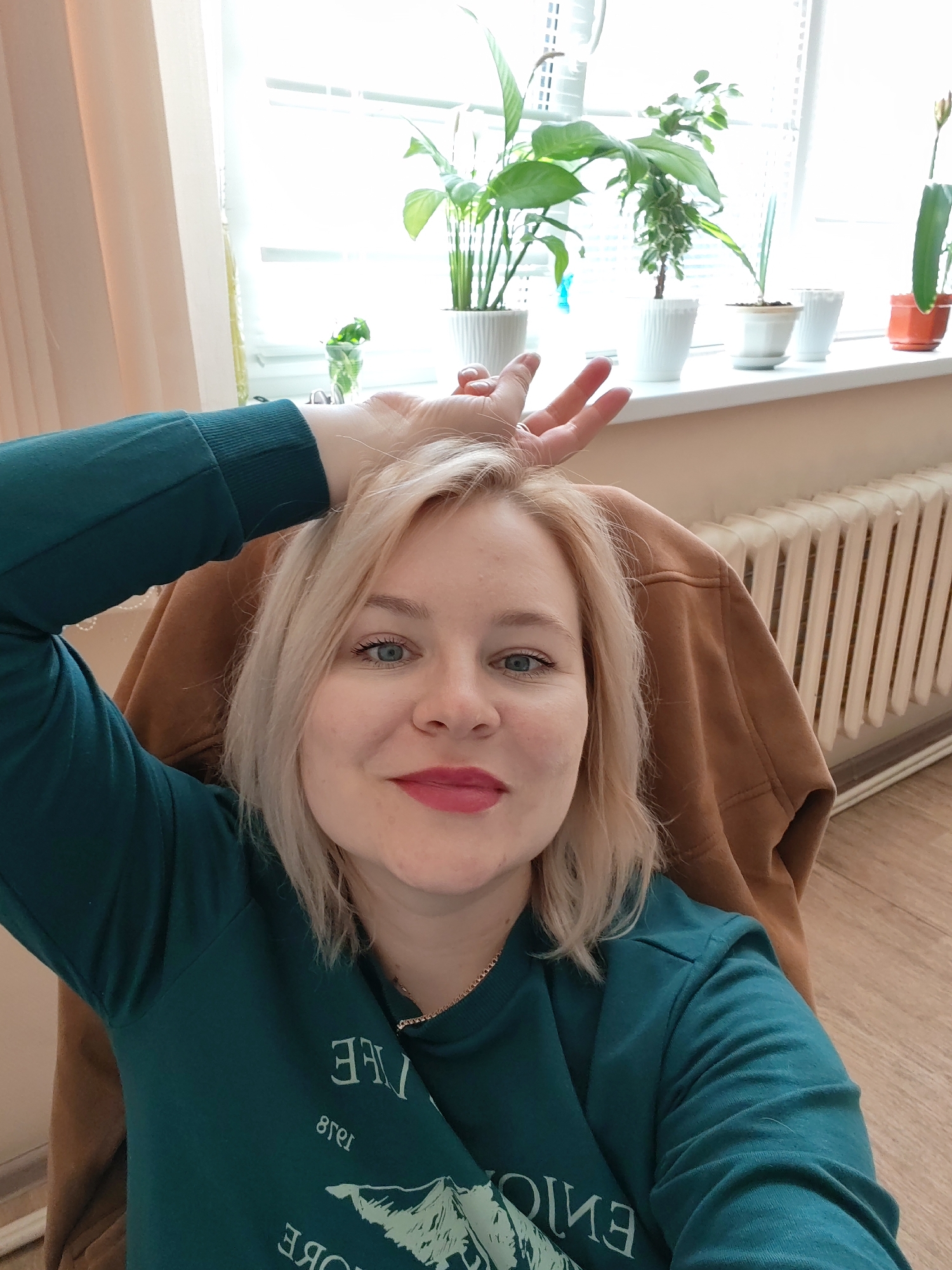 A fair-skinned woman with shoulder-length blonde hair and blue-green eyes is captured in a selfie indoors, likely in Izhevsk, Russia. She is looking directly at the camera with a gentle, pleasant smile. Her left arm is raised, with her hand near her head, in a relaxed pose. She wears a dark teal long-sleeved top that features a light-colored, mirrored design and text on the front. Parts of the text are visible as "FREE" and "LIFE," along with what appears to be a mountain graphic. A thin gold-colored necklace is visible around her neck. She is seated, with a brown hooded garment providing a backrest.
The background shows a bright, daytime interior. A large window dominates the upper portion, fitted with white horizontal blinds and a white windowsill. A collection of diverse potted plants, including a peace lily, succulents, and other greenery, are arranged on the windowsill in white and one orange pot. To the woman's right, a tall, light-colored radiator with vertical sections is prominent. Beige or light brown curtains frame the left side of the window. The flooring appears to be light-colored wood. The scene conveys a relaxed and comfortable ambiance.