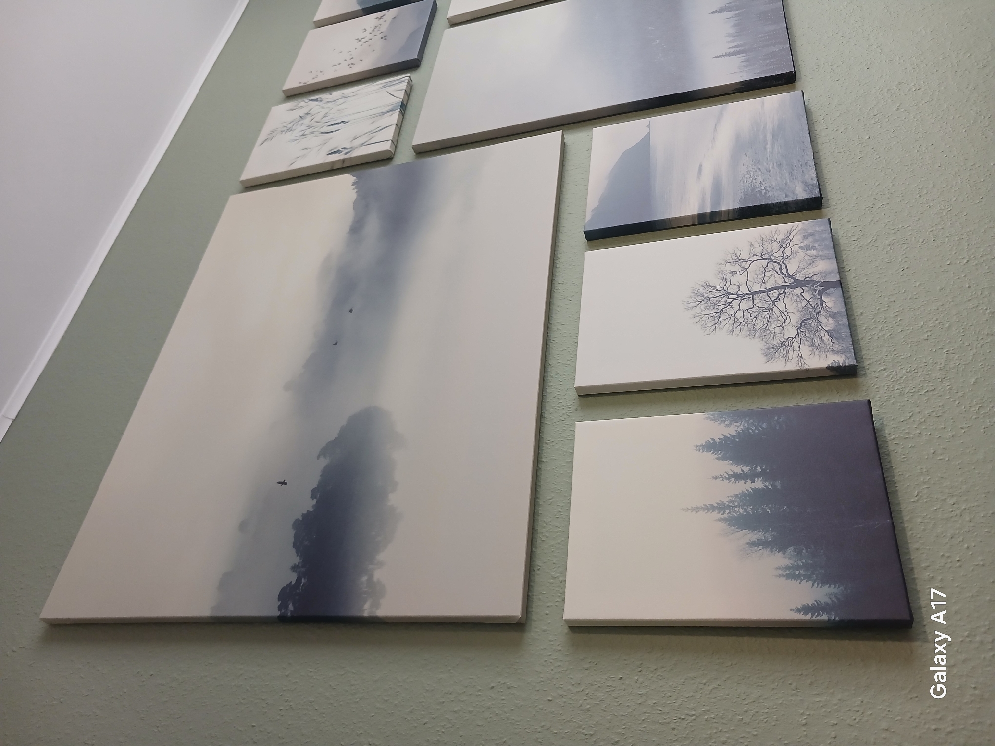 A close-up, slightly angled shot reveals a collection of framed artworks hanging on a light green, textured wall. The artworks, primarily square or rectangular in shape, feature grayscale landscape scenes depicting elements such as trees, mountains, and sky. The largest artwork dominates the left side of the composition and showcases a cloudy, atmospheric vista with a solitary bird in flight. The other artworks are smaller and arranged in a grid-like fashion to the right of the large piece. The overall aesthetic is minimalist and serene. A camera watermark, "Galaxy A17," is present in the lower right corner of the image. The location cannot be determined.