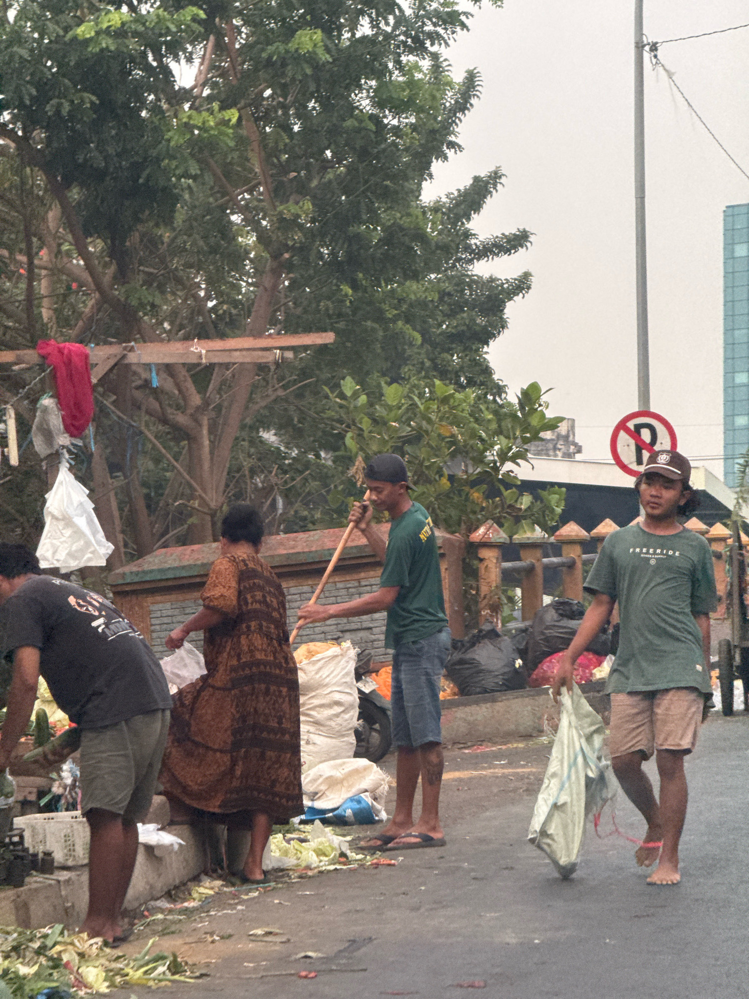 The image depicts a street scene in Surabaya, Indonesia, with several people engaged in activity around piles of garbage and refuse. In the foreground, a person in a dark t-shirt and light-colored shorts is bent over, interacting with debris. To their right, a person wearing a brown patterned dress is holding a white plastic bag. Further right, a person in a green t-shirt and dark shorts is holding a broom and appears to be sweeping or cleaning. Behind them, another person in a green t-shirt and blue denim shorts is also present. Various garbage bags, including black and white sacks, are visible, along with scattered organic waste, possibly from a market. In the background, trees line the street, and a modern building can be seen in the distance. A "P" parking sign with a red circle and diagonal line is also visible. The sky is overcast, suggesting a potentially humid or hazy day.