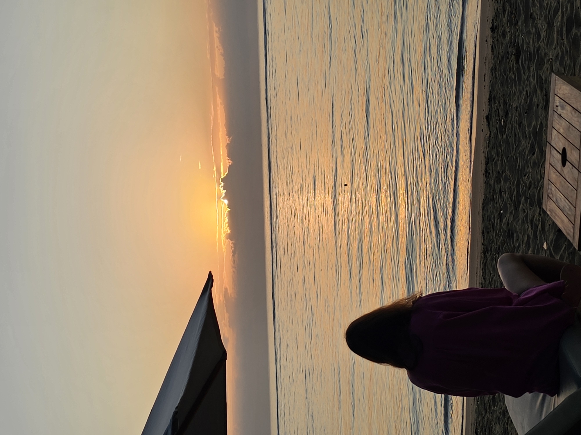 A person is seated on a lounge chair on a sandy beach, facing the ocean. The sun is setting, casting a warm, orange glow over the water and sky. Clouds are visible on the horizon, partially obscuring the sun. A beach umbrella is positioned to the left of the frame, and a small wooden table is in the foreground. The scene is peaceful and serene. This image was taken in Denpasar, Indonesia.