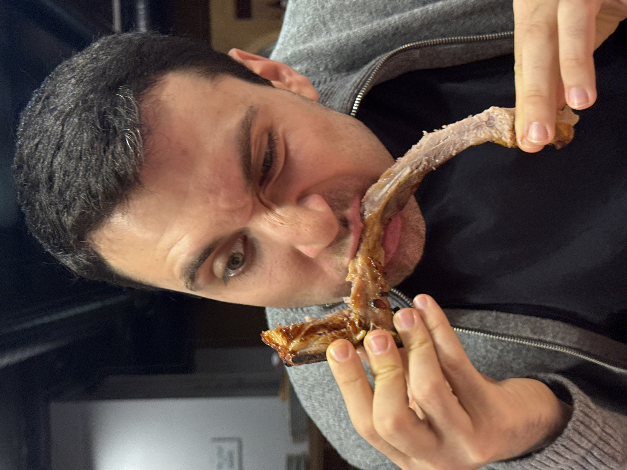 A man is shown in a close-up shot, eating meat off a bone. He's wearing a gray zip-up hoodie over a dark shirt. His dark hair is short, and he is looking to his left. The location and city are not discernible from the given image.
