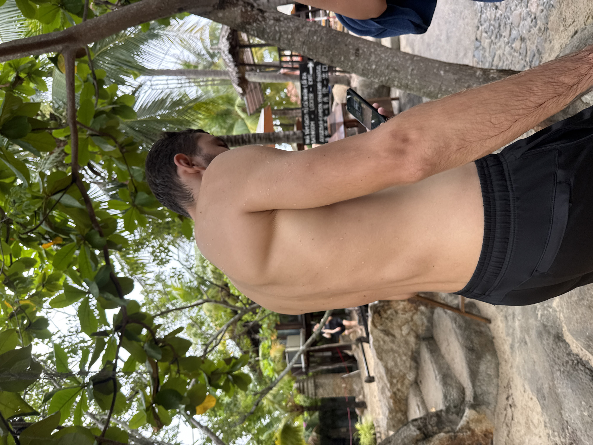A shirtless man with short dark hair, wearing black shorts, stands outdoors, looking down at a smartphone in his right hand. He is holding the phone near his chest, and the screen is illuminated. The man's skin is wet with droplets visible. In the background, there are lush green trees and palm trees, suggesting a tropical environment. Further in the background, a traditional thatched-roof building and some stone structures are visible, possibly indicating a resort or tourist area. A sign with text in Vietnamese and English is also present, mentioning "BATHROOM," "LOCKER," and "EXIT." The ground appears to be sandy and rocky, with stone steps leading up to the left. The overall scene depicts a relaxed outdoor setting, likely in Nha Trang, Vietnam, given the presence of Vietnamese text.