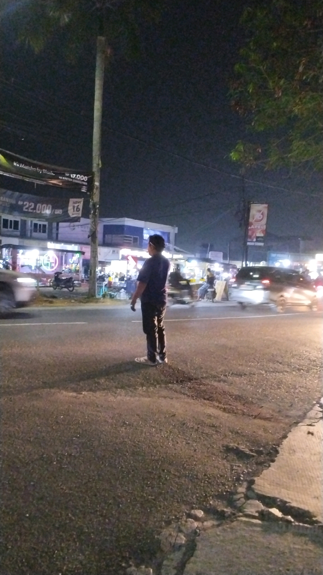 A full-body shot captures a young man standing in the middle of a road at night in Canggu, Indonesia. He is wearing a dark blue t-shirt and dark pants, with white shoes. The man stands with his back to the camera, facing away from the viewer, looking towards a street filled with shops and parked vehicles. To the right of the man, a white van is visible driving down the road. The road itself appears to be paved and shows some wear and tear, with cracks and uneven surfaces. In the background, shops and street vendors are illuminated by bright lights, creating a vibrant and busy atmosphere. Trees and power lines are also visible, suggesting an urban or semi-urban setting. The sky is dark, indicating nighttime.