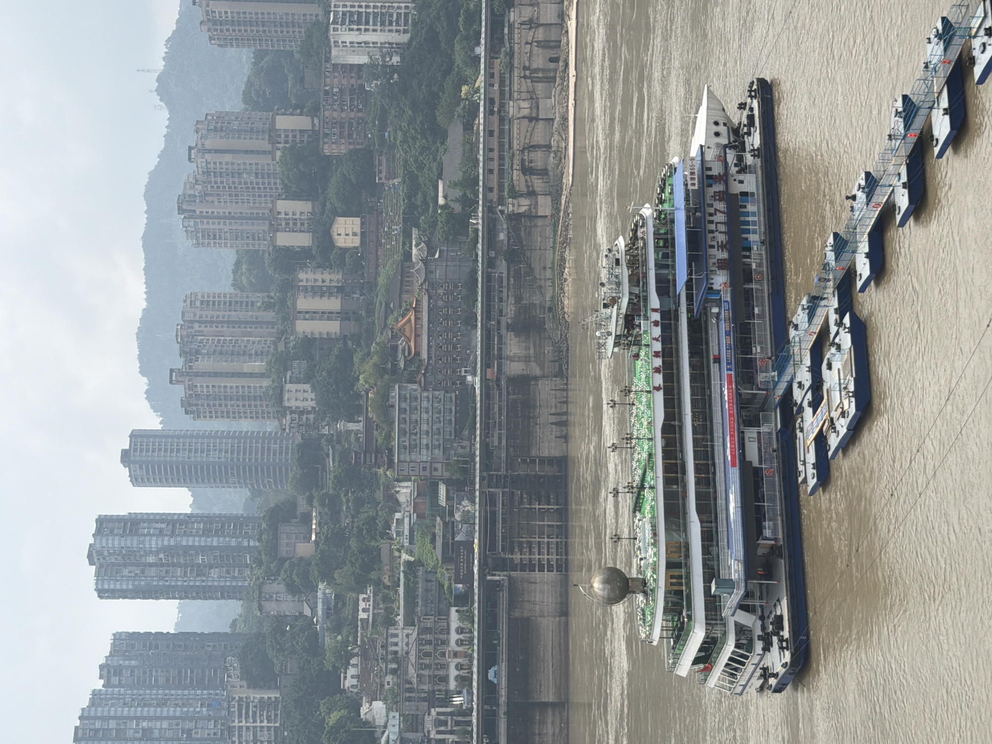 In the Yuzhong District of China, a large, multi-story passenger ship with white and green decks is docked on a wide, murky river. The ship has a large spherical ornament on its top deck and is connected to a series of floating docks via gangplanks. Several smaller, blue and white boats are attached to these docks. In the background, a densely packed cityscape rises up a steep hill. Numerous tall, modern apartment buildings and older, traditional structures are visible, interspersed with lush green vegetation. A substantial concrete embankment with stair-like structures runs along the riverfront, separating the water from the city. The sky is overcast, casting a diffused light on the scene.