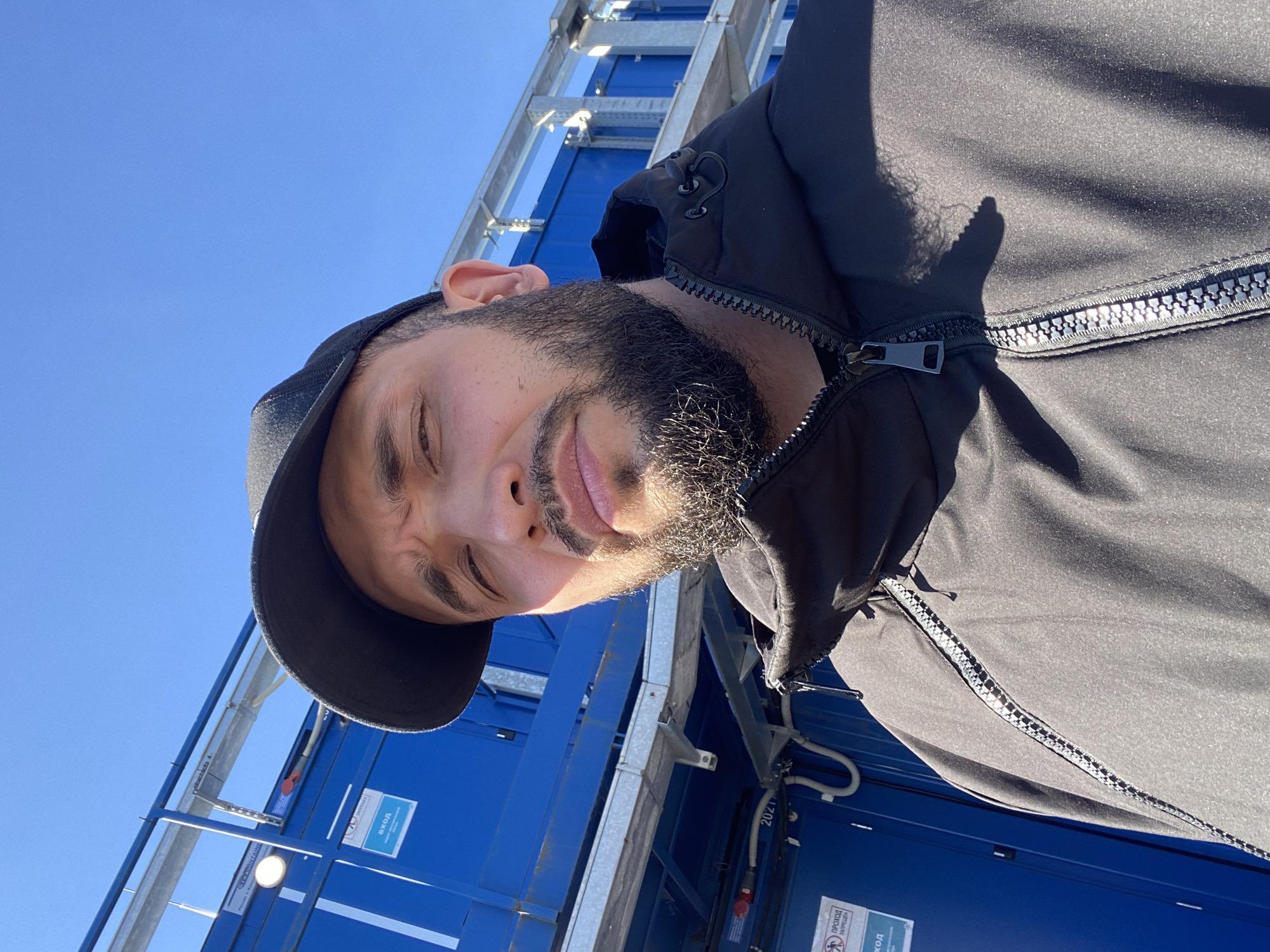 A man with dark hair and a beard wearing a black cap and black jacket poses in front of a blue structure in Koskolovo, Russia. The structure appears to be composed of stacked units, and is framed by metal supports. Signage with Russian text is visible on the blue units. The sky is clear and blue. The man's face is partially shadowed.