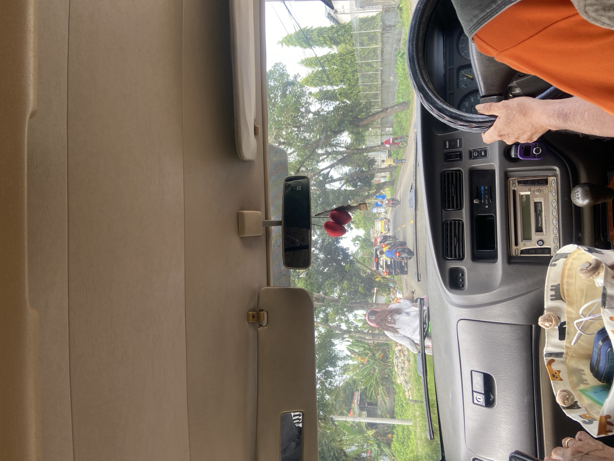 The image is taken from the interior of a vehicle in Palopo, Indonesia, looking forward through the windshield. The perspective is from the passenger seat or the driver's perspective, showing the dashboard, steering wheel, and a portion of the driver's arm wearing an orange shirt. Outside the vehicle, a street scene unfolds with a clear day. Several people are visible, including a person in the foreground on a motorcycle wearing a white hoodie and a pink helmet, facing away from the camera. Further down the road, other motorcycles and bicycles are in motion, along with some cars. The environment appears to be a typical roadside in a city, with trees lining the street, some houses or buildings visible in the background, and a fence. The road itself has markings, indicating it's a functioning street. A decorative item, possibly two red hearts, hangs from the rearview mirror inside the vehicle.